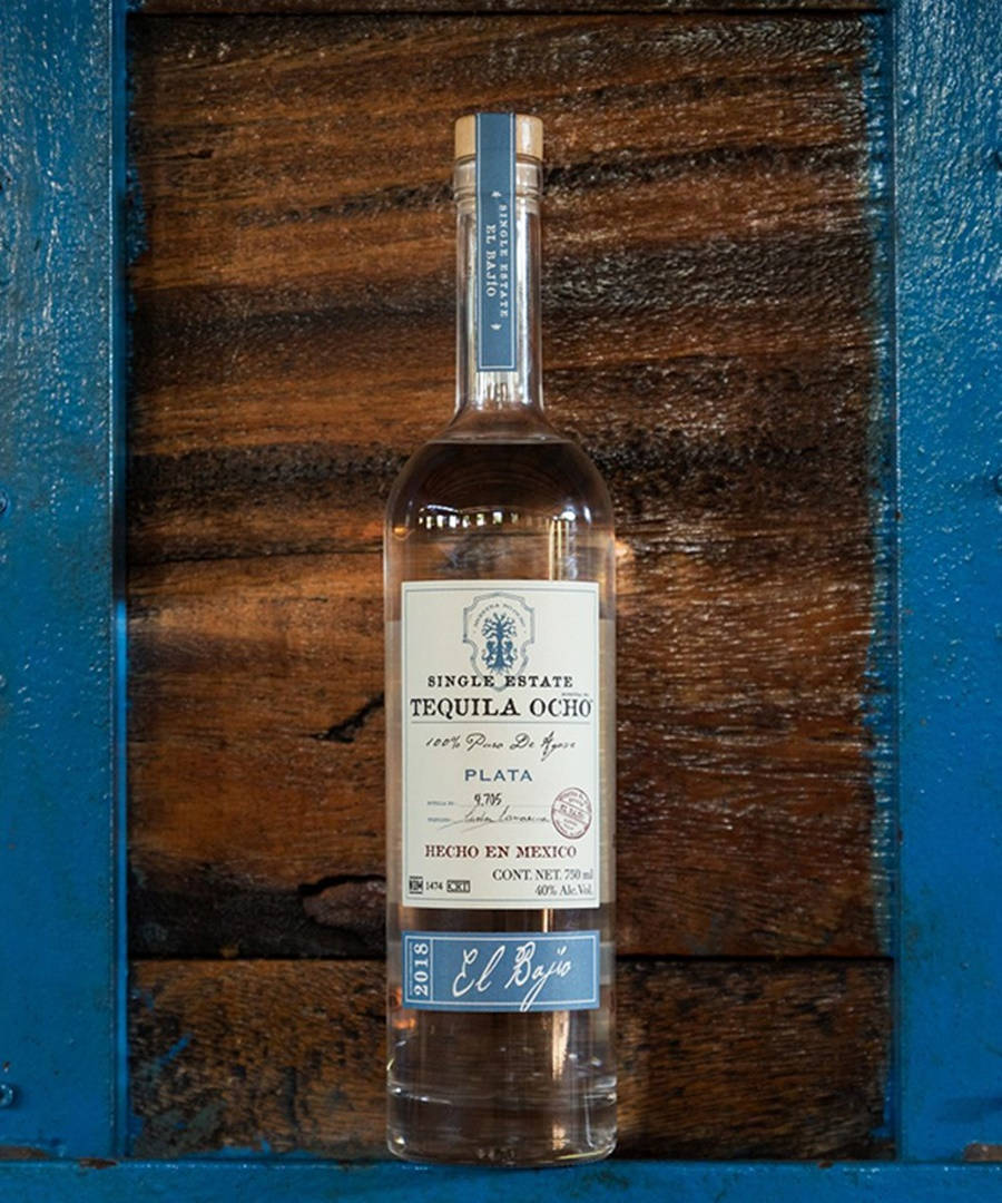 Tequila Ocho 奢華酒品 壁紙 — 一瓶清澈的 Single Estate Tequila Ocho Plata 置中於質樸的木質背景前，並以鮮豔的藍色油漆框起。