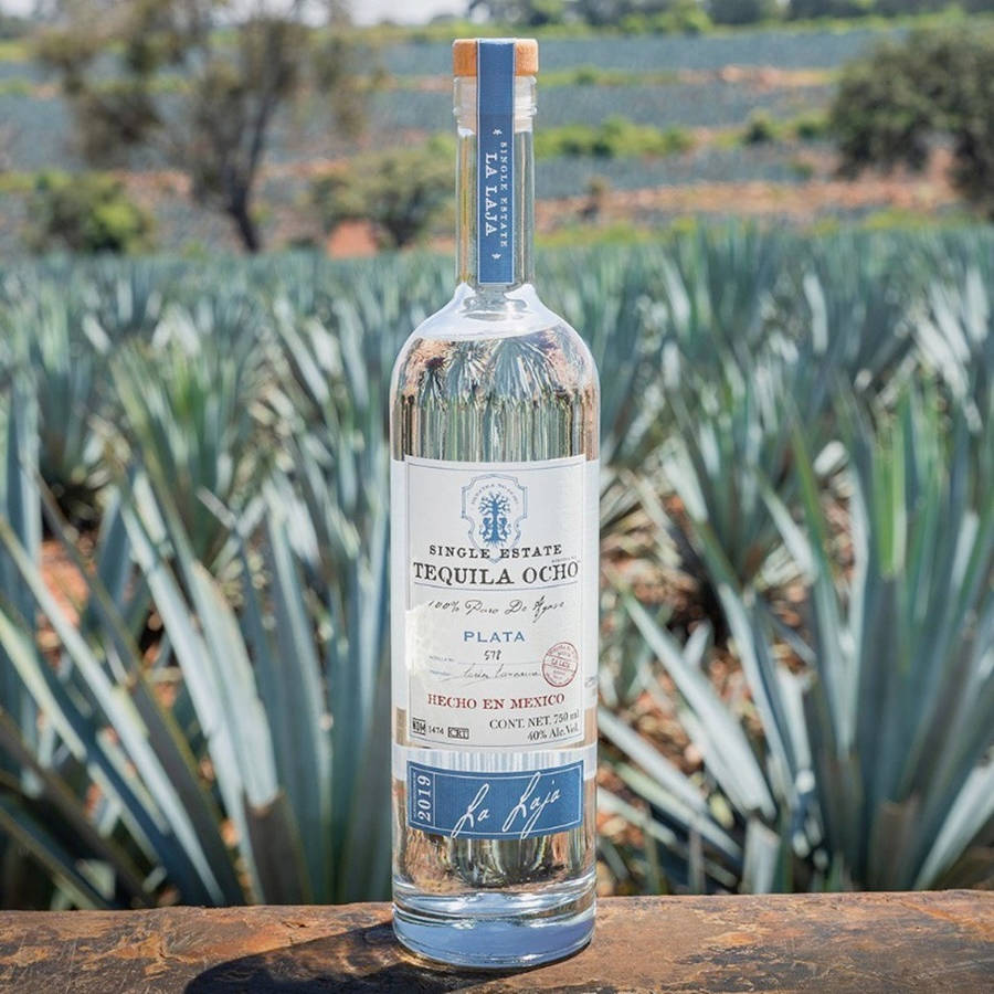 藍標 Tequila Ocho 龍舌蘭田 壁紙 — 一個透明嘅龍舌蘭酒樽，標籤寫住 'Single Estate Tequila Ocho'，放在木質表面上，背景有龍舌蘭植物。