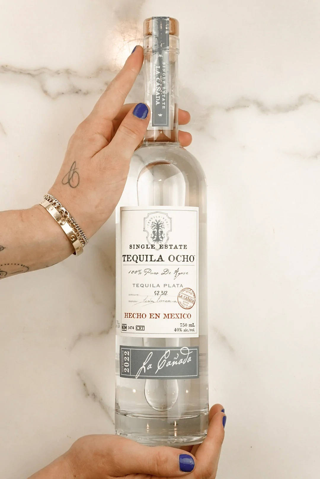 Tequila Ocho 酒品手模展示 壁紙 — 一隻塗有藍色指甲油的手，在白色大理石背景前拿著一瓶透明的Tequila Ocho。