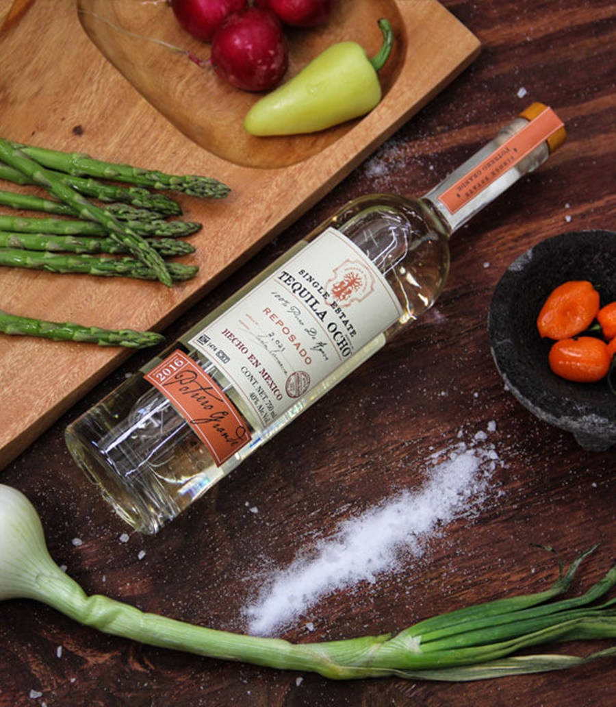 Tequila Ocho 最佳酒品 壁紙 — 一瓶龍舌蘭酒雷波薩多放在深色木桌上，周圍是新鮮的蘆筍、蘿蔔和辣椒。