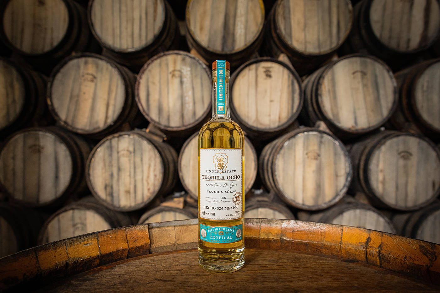 Tequila Ocho 烈酒歷史風貌 壁紙 — 一瓶 Single Estate Tequila Ocho 放在木桶上，背景是一排排陳年的木桶。
