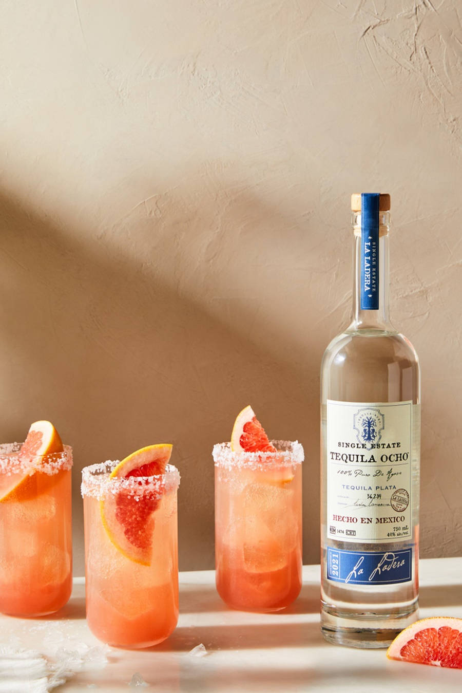 藍標 Tequila Ocho 酒瓶與酒杯 壁紙 — 三杯充滿活力的粉紅色龍舌蘭雞尾酒，配上西柚裝飾和鹽邊，旁邊放著一瓶La Patrera龍舌蘭酒，置於有紋理的米色表面上。