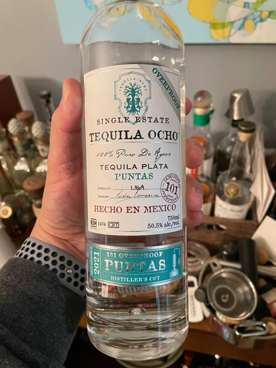 白標 Tequila Ocho Plata 酒品 壁紙 — 一隻手拿著一瓶透明的101 proof Tequila Ocho，背景是酒瓶和酒吧工具。