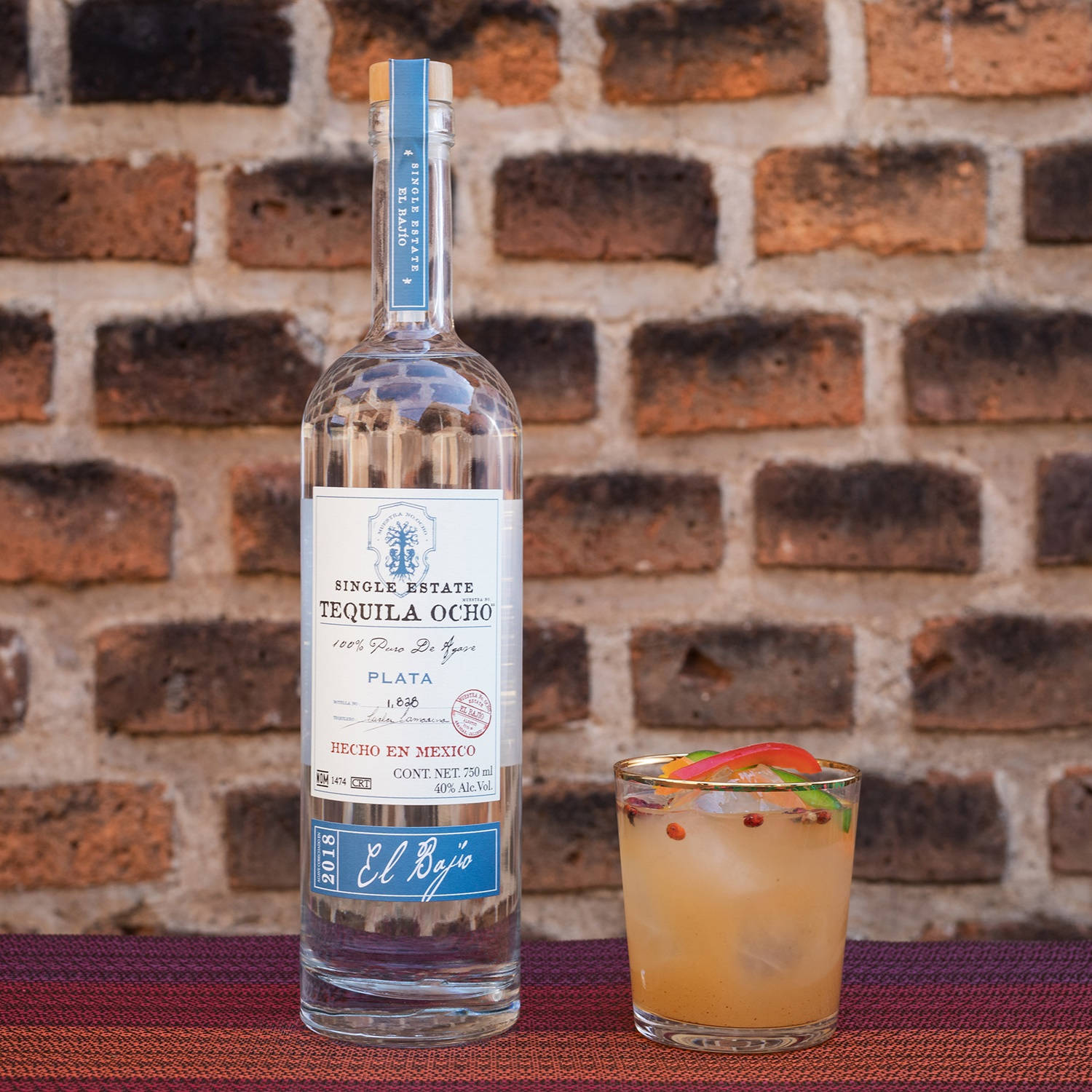 Tequila Ocho Plata 龍舌蘭酒搭配橙色飲品 壁紙 — 一瓶 El Bajo Tequila Plata 放在質感地毯上，旁邊是一杯以辣椒和青檸裝飾的鮮艷雞尾酒，背景是質樸的磚牆。