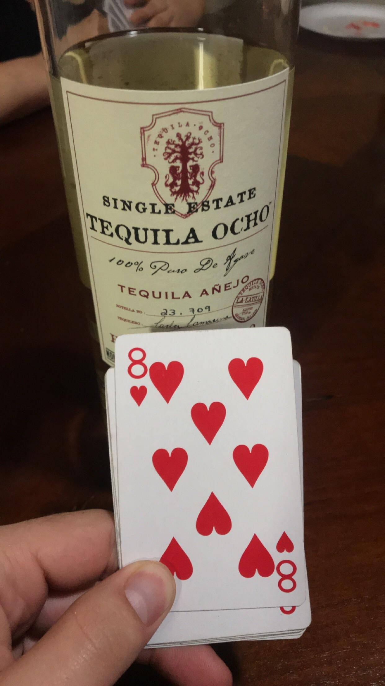 Tequila Ocho 酒類紙牌遊戲 壁紙 — 一隻手在木桌上，拿著一張紅心8牌，放在一瓶龍舌蘭酒前面。