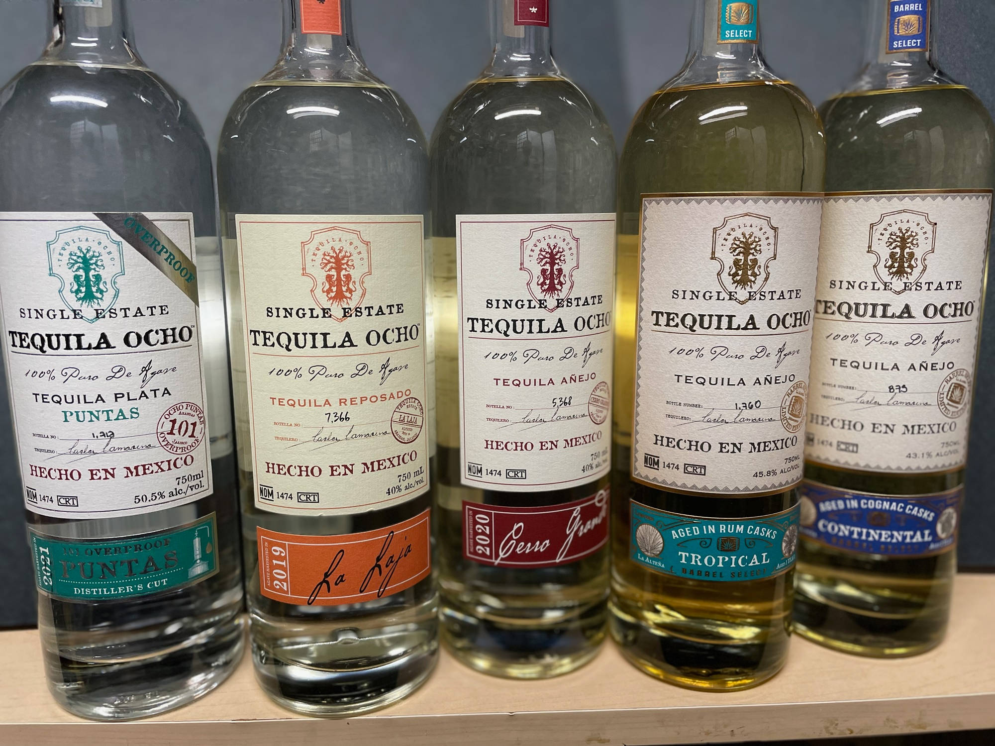 Tequila Ocho 陳釀桶選系列 壁紙 — 五個標有標籤的龍舌蘭酒瓶，年齡和風格各異，整齊排列在木架上，帶有透明玻璃和彩色標籤。
