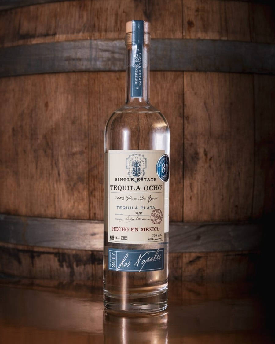 一瓶標有 Single Estate Tequila Ocho 的透明龍舌蘭酒瓶放在木桶前的木質表面上。
