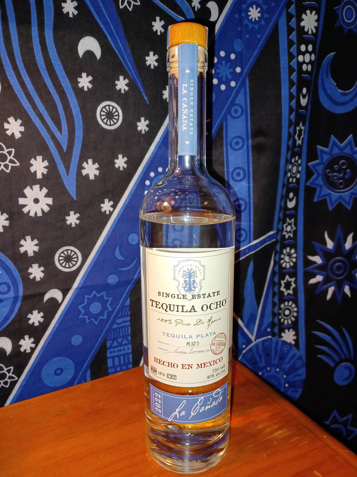 一個Tequila Ocho酒瓶放在木頭上，背景是藍色圖案布。