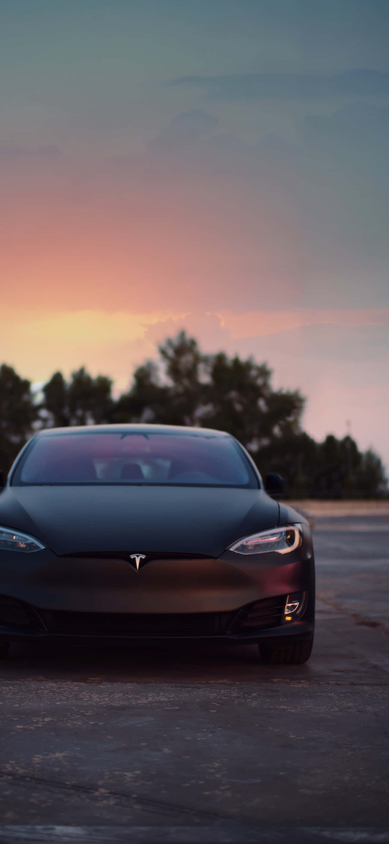 享受無線世界的自由 —— Tesla iPhone 壁紙 — 一輛光滑的啞光黑色Tesla停在瀝青路上，背景是充滿活力的日落天空和剪影樹木。