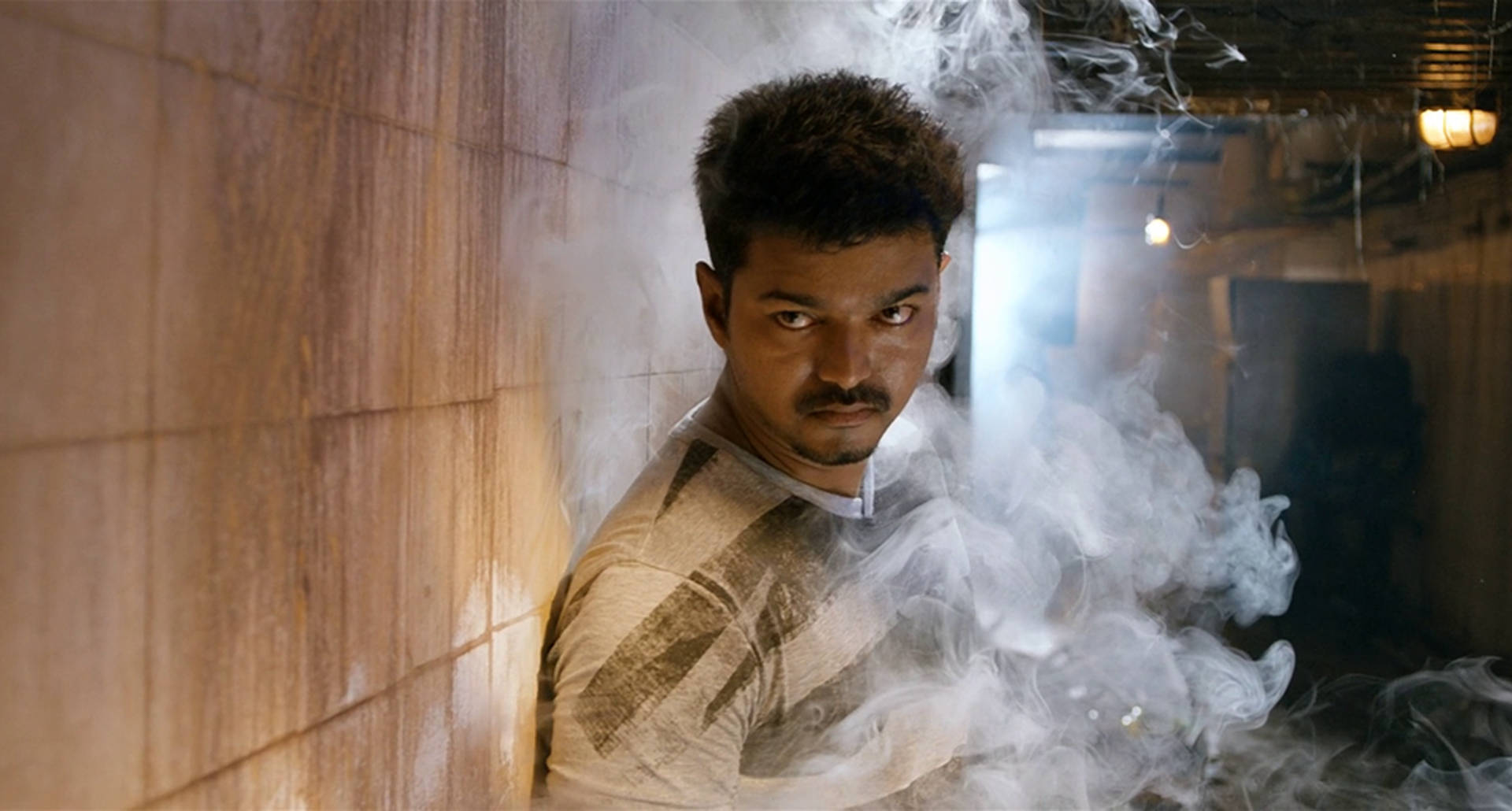 Thalapathy HD電影場景 壁紙 — 一個眼神凌厲、留著鬍子的男人，站在煙霧瀰漫、燈光昏暗的小巷中，背靠瓷磚牆。