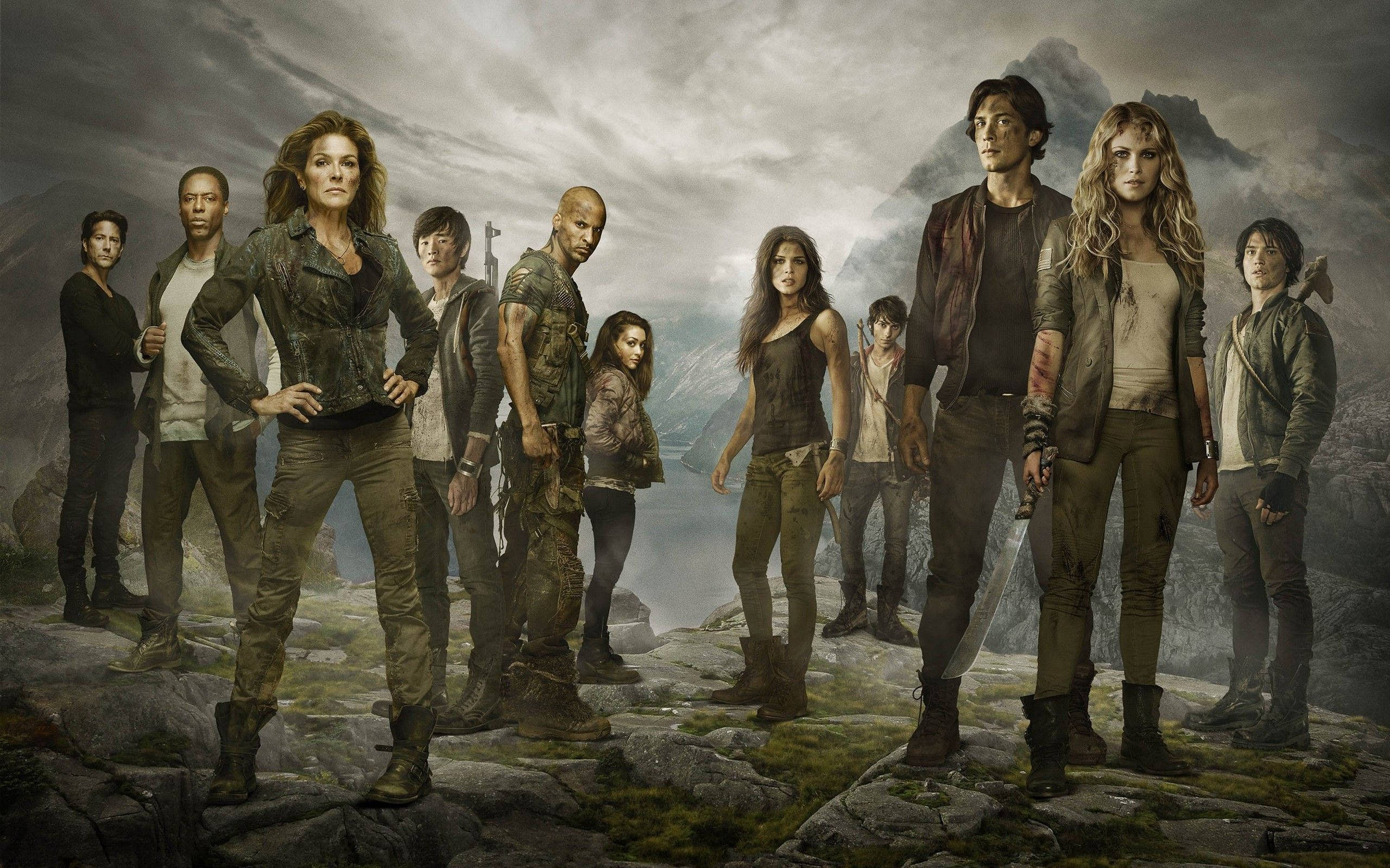 《The 100》女性角色阿比蓋爾·格里芬 壁紙 — 一群強悍的倖存者挑釁地站在長滿苔蘚的懸崖上，背景是暴風雨的山脈。