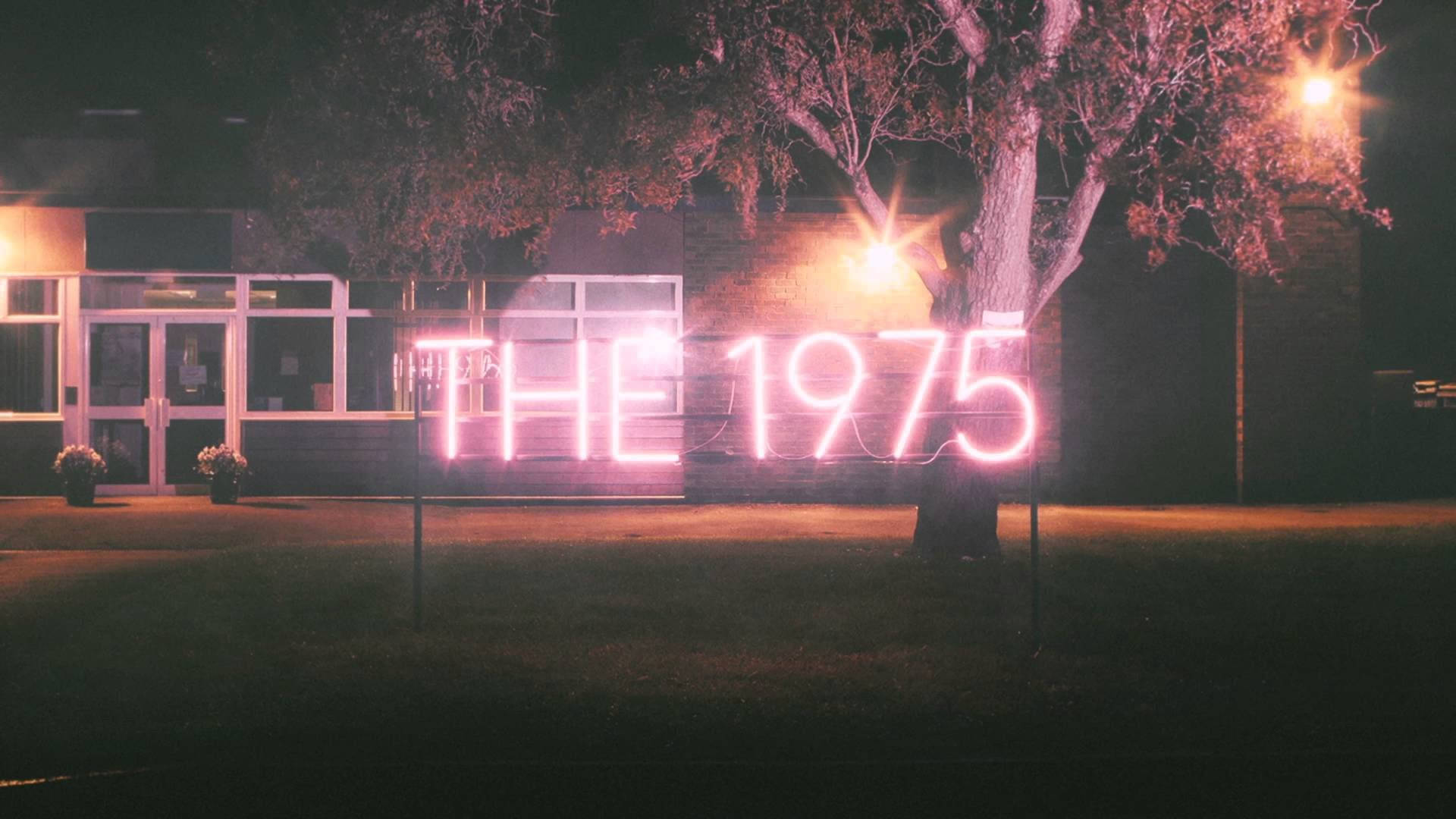 The 1975 粉紅霓虹燈招牌 壁紙 — 夜晚，一棵樹下的建築物前，有一個發光的粉紅色霓虹燈牌，上面寫著 THE 1975。