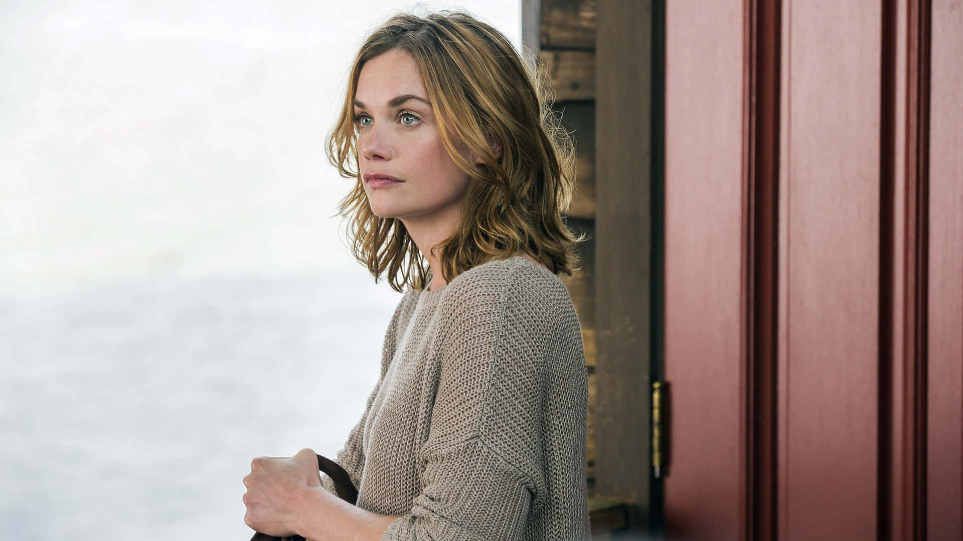 《The Affair》海邊沉思女子 壁紙 — 一位身穿米色針織毛衣的女性站在紅色門旁，若有所思地凝視著模糊的水景。