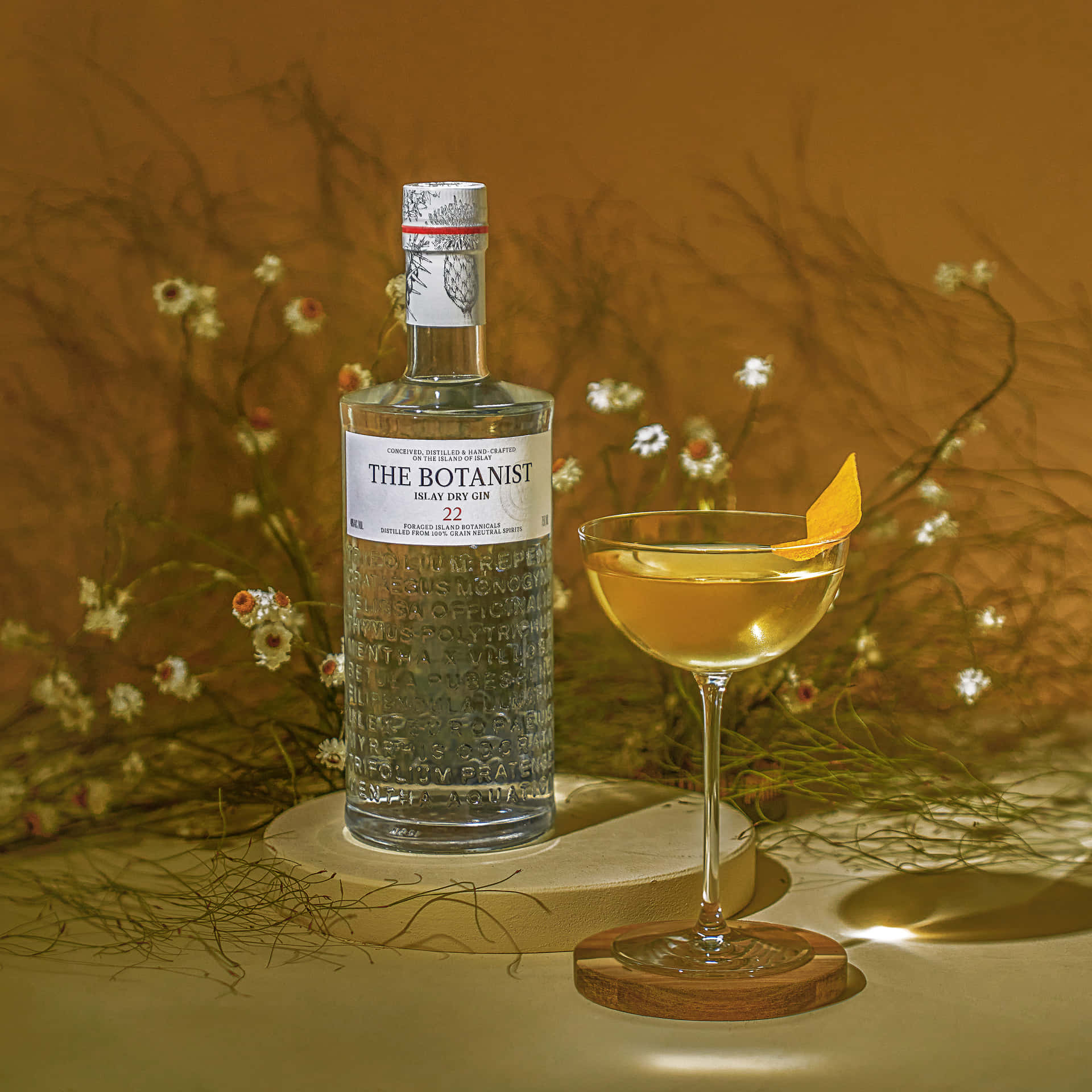 一瓶 The Botanist 氈酒和一杯橙裝飾雞尾酒，放在木杯墊上，伴有乾花。