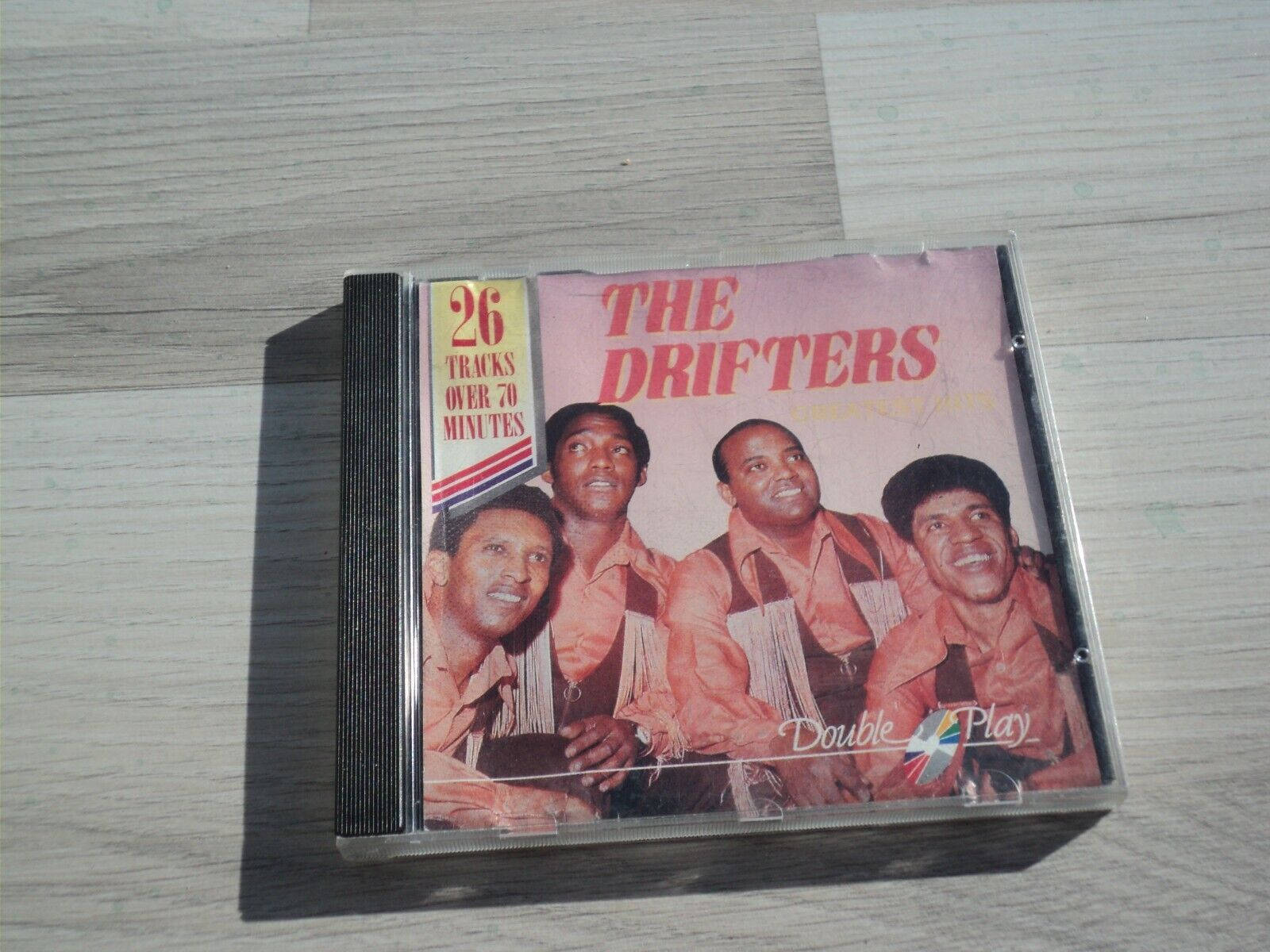 The Drifters CD 專輯封面 壁紙 — 一個The Drifters的復古CD盒放在淺色木質表面上，顯示了樂隊的笑臉和'Double Play'品牌標誌。