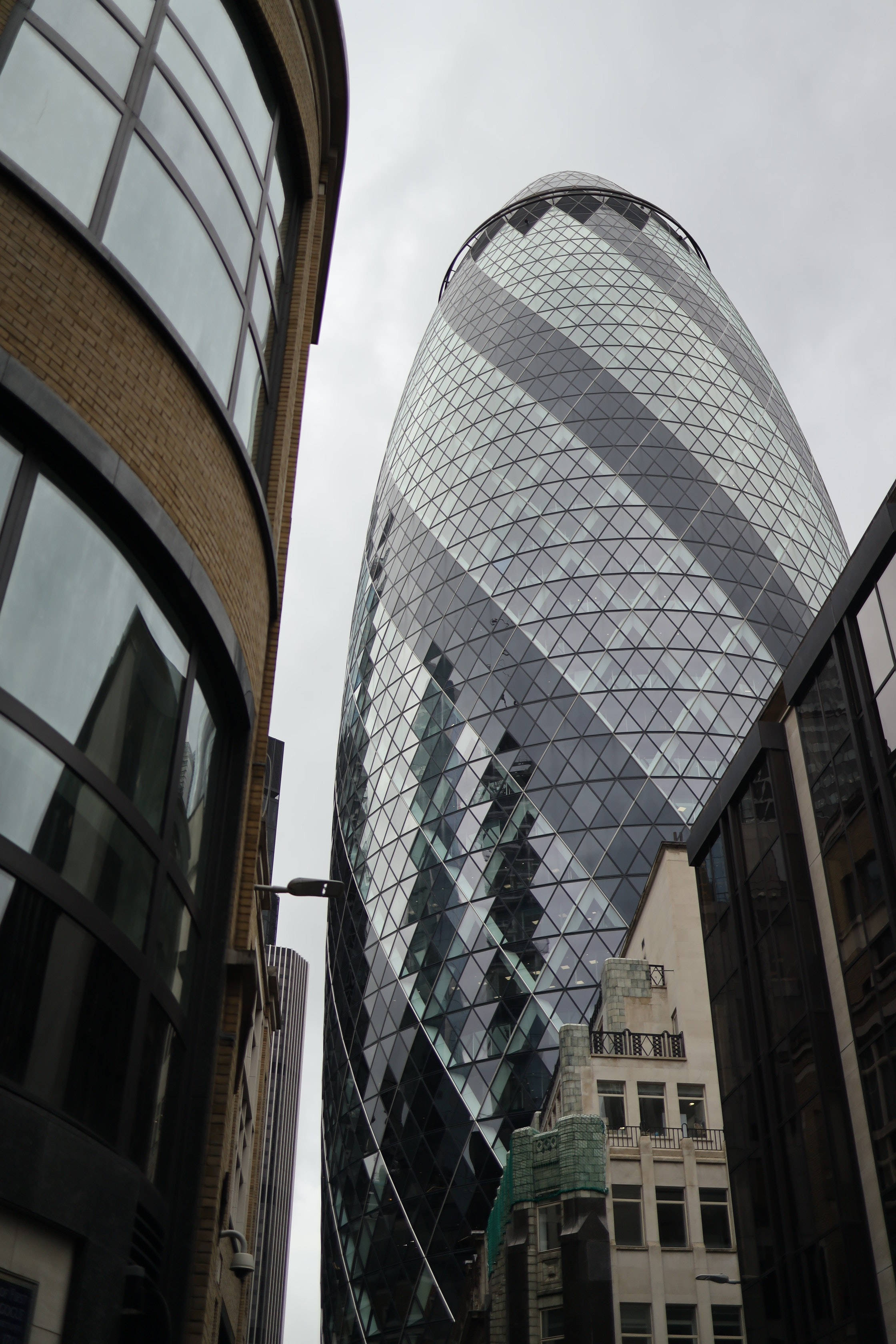 The Gherkin與鄰近建築 壁紙 — 從低角度觀看標誌性的Gherkin摩天大樓，其玻璃外牆反射灰色雲層，周圍環繞著城市建築。