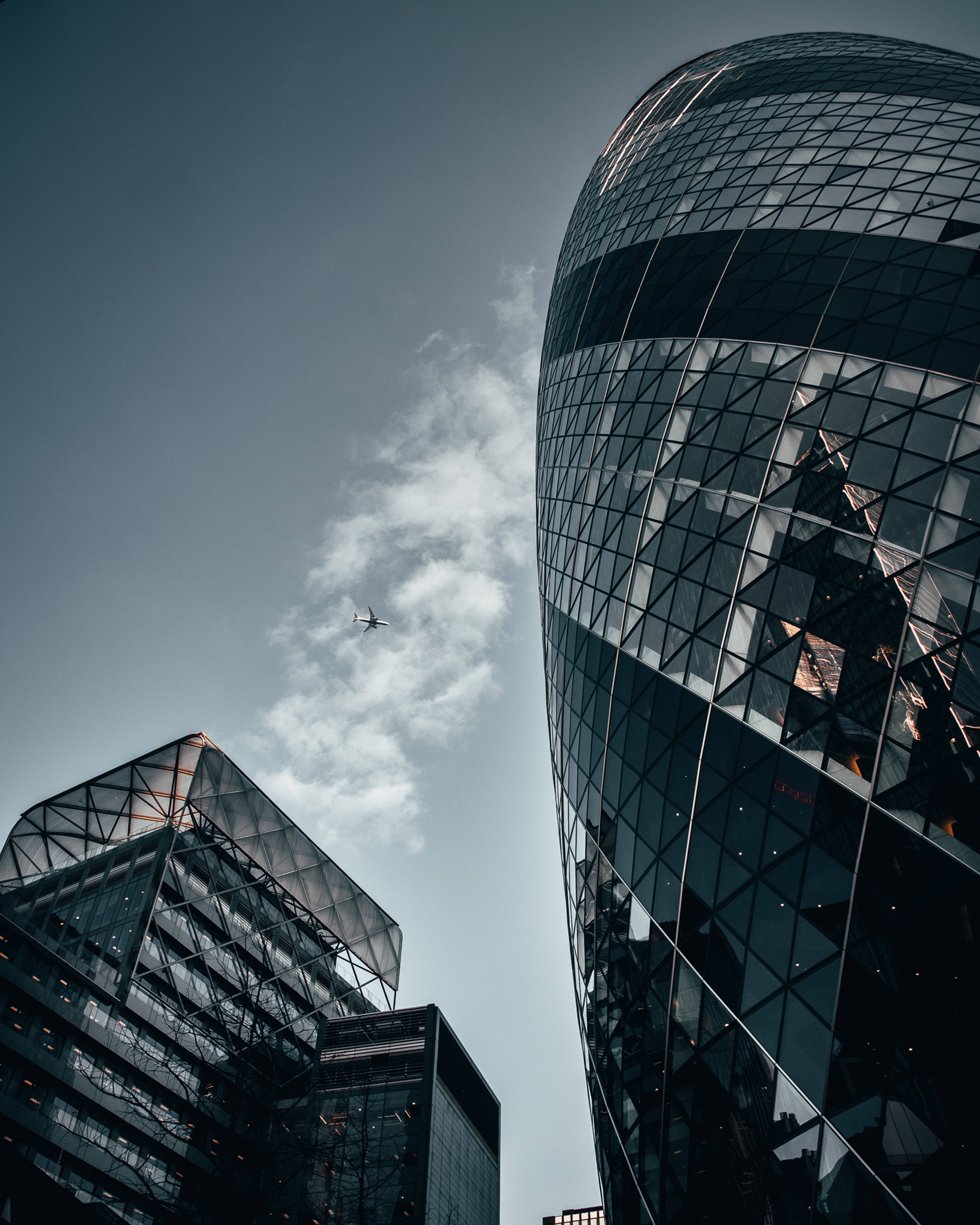 The Gherkin 現代高樓建築 壁紙 — 從低角度觀看現代玻璃摩天大樓，天空多雲，遠處有飛機飛過。