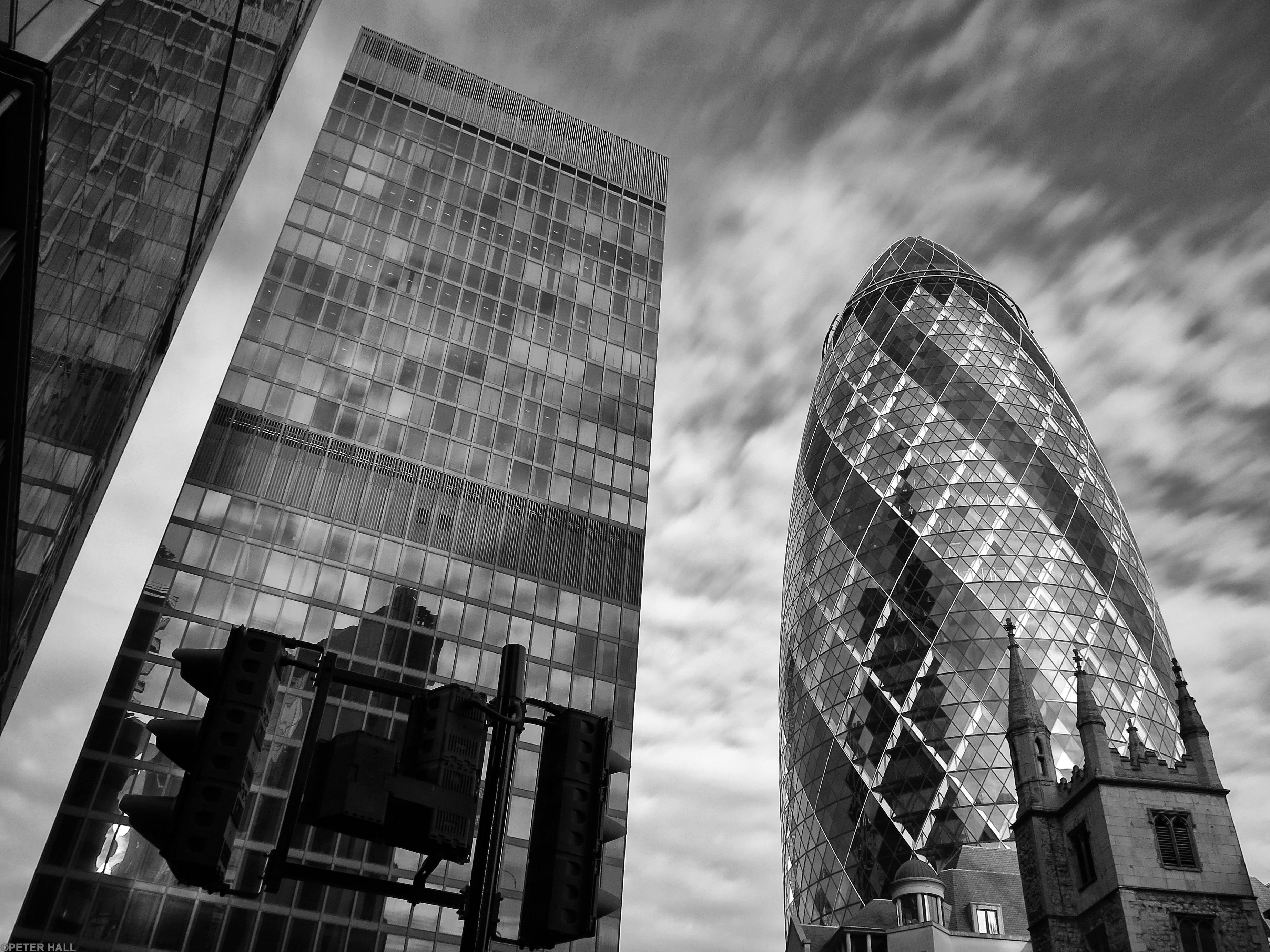 The Gherkin 黑白攝影 壁紙 — 一張戲劇性的黑白低角度照片，拍攝了倫敦標誌性的小黃瓜摩天大樓旁邊的一座傳統教堂，背景是陰天。