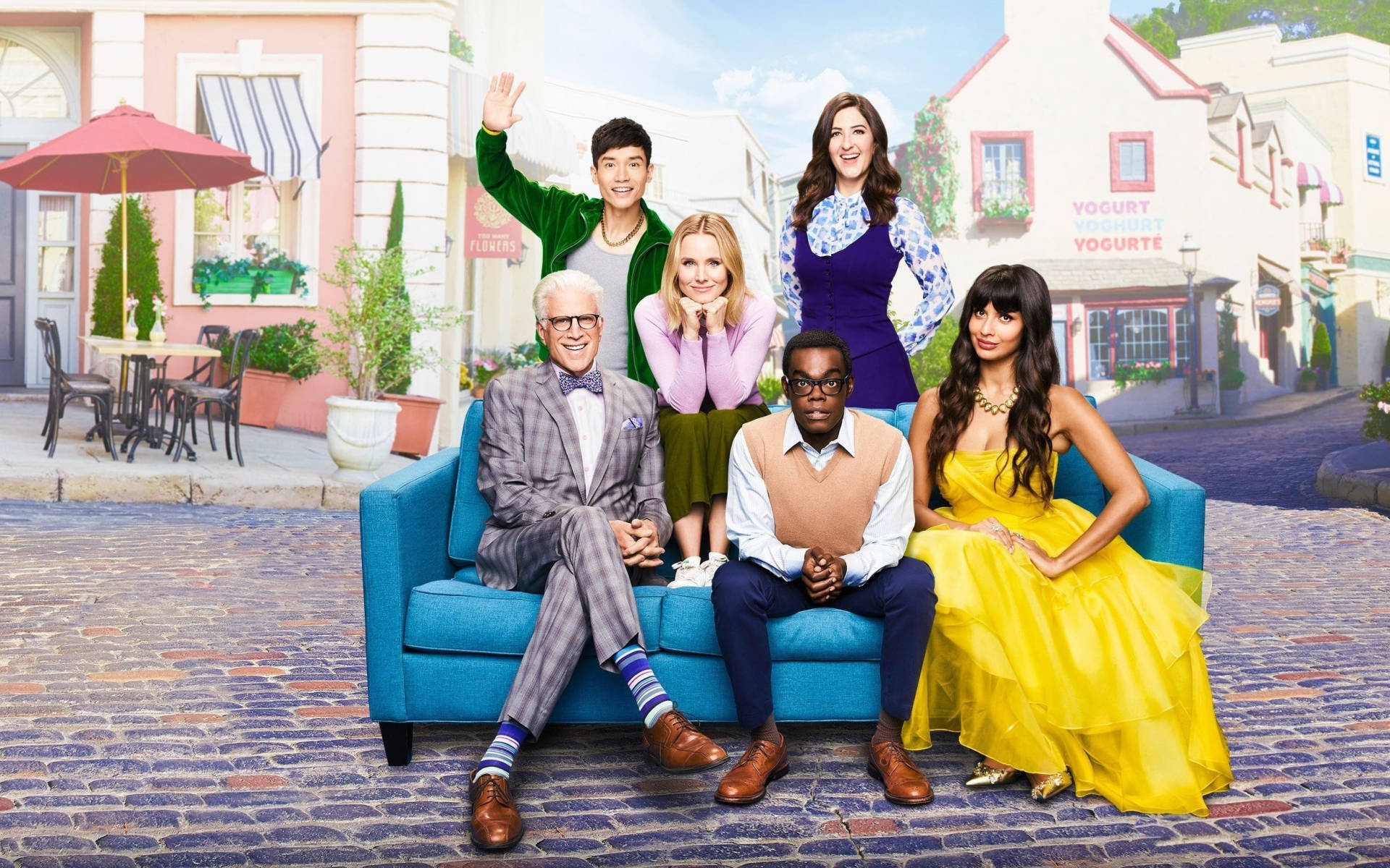 《The Good Place》主要演員於時尚藍沙發上合影 壁紙 — 六位微笑的演員在色彩繽紛的歐洲風格街景中，在明亮的藍色沙發上擺姿勢，背景有鵝卵石和咖啡館遮陽傘。