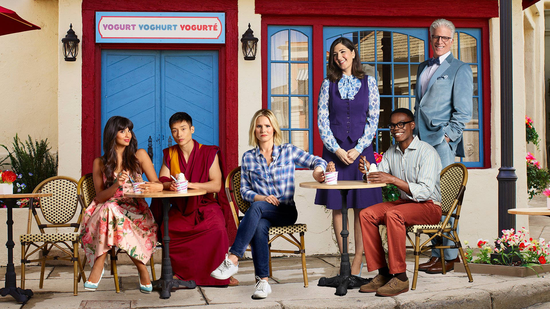 《The Good Place》角色於戶外餐廳休閒聚會 壁紙 — 六位不同人士在戶外咖啡桌享用乳酪，背景是一座色彩繽紛的建築物，有藍色門和紅色飾邊。