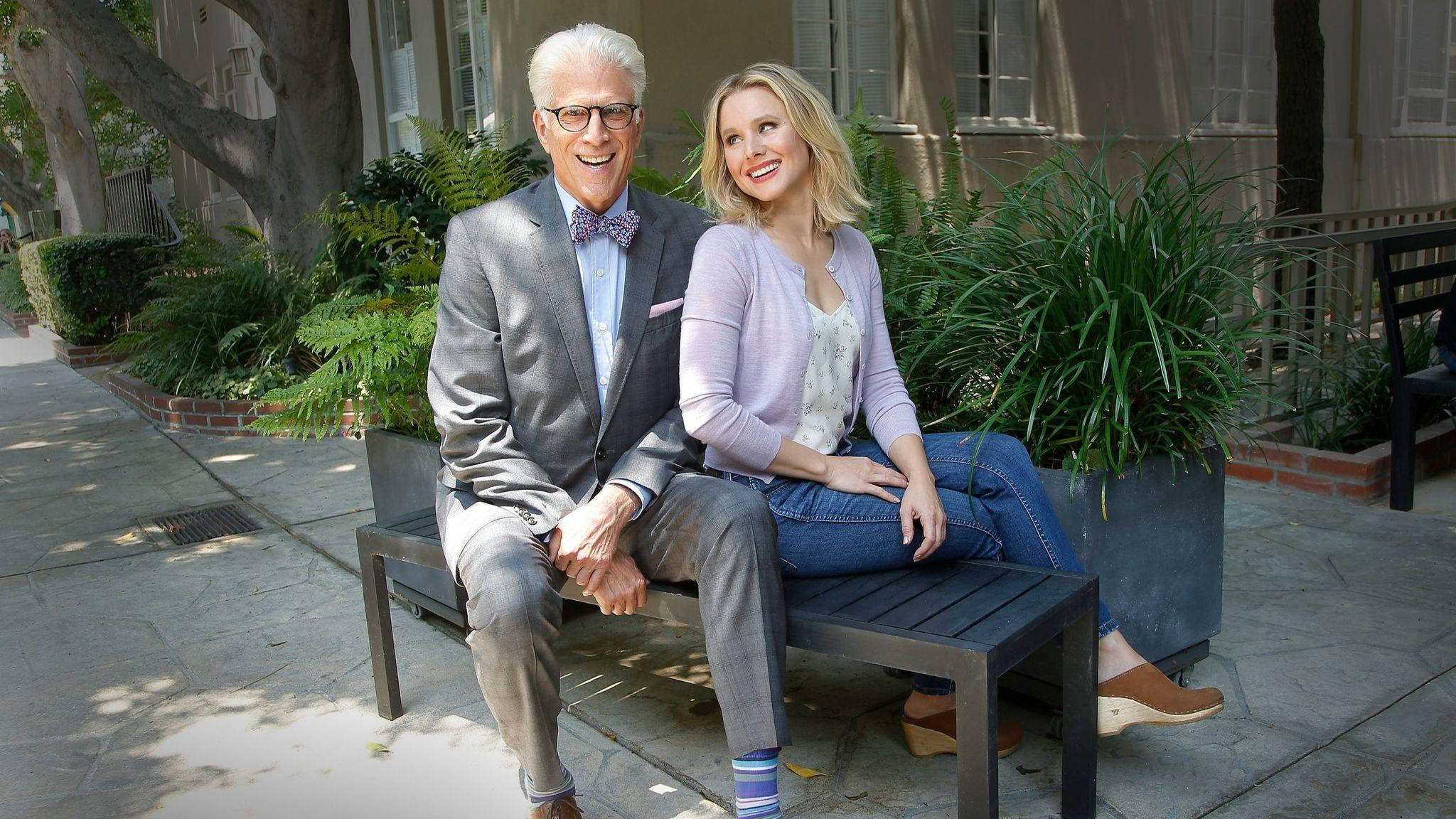 《The Good Place》邁克爾與愛蓮娜 壁紙 — 一位開朗的長者與年輕女士坐在陽光普照的庭院長椅上，四周綠樹成蔭，兩人面帶微笑。