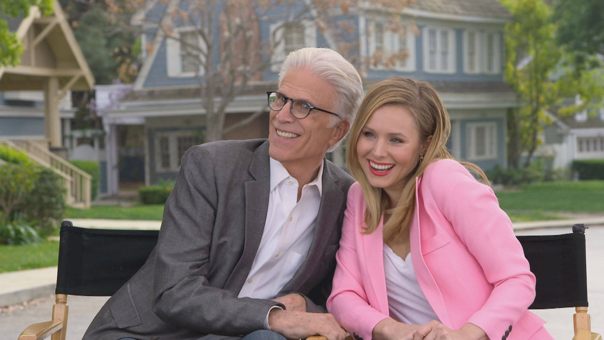 《The Good Place》邁克爾與愛蓮娜拍照時刻 壁紙 — 一位年長男士和一位年輕女士在藍色房屋和綠色草坪的郊區社區中，坐在導演椅上一起微笑。