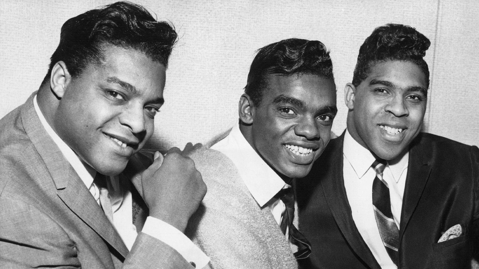 The Isley Brothers英國巡迴演出照片 壁紙 — 三位微笑的黑人男士穿著俐落的西裝，在經典黑白肖像中以樸素背景自信地擺姿勢。