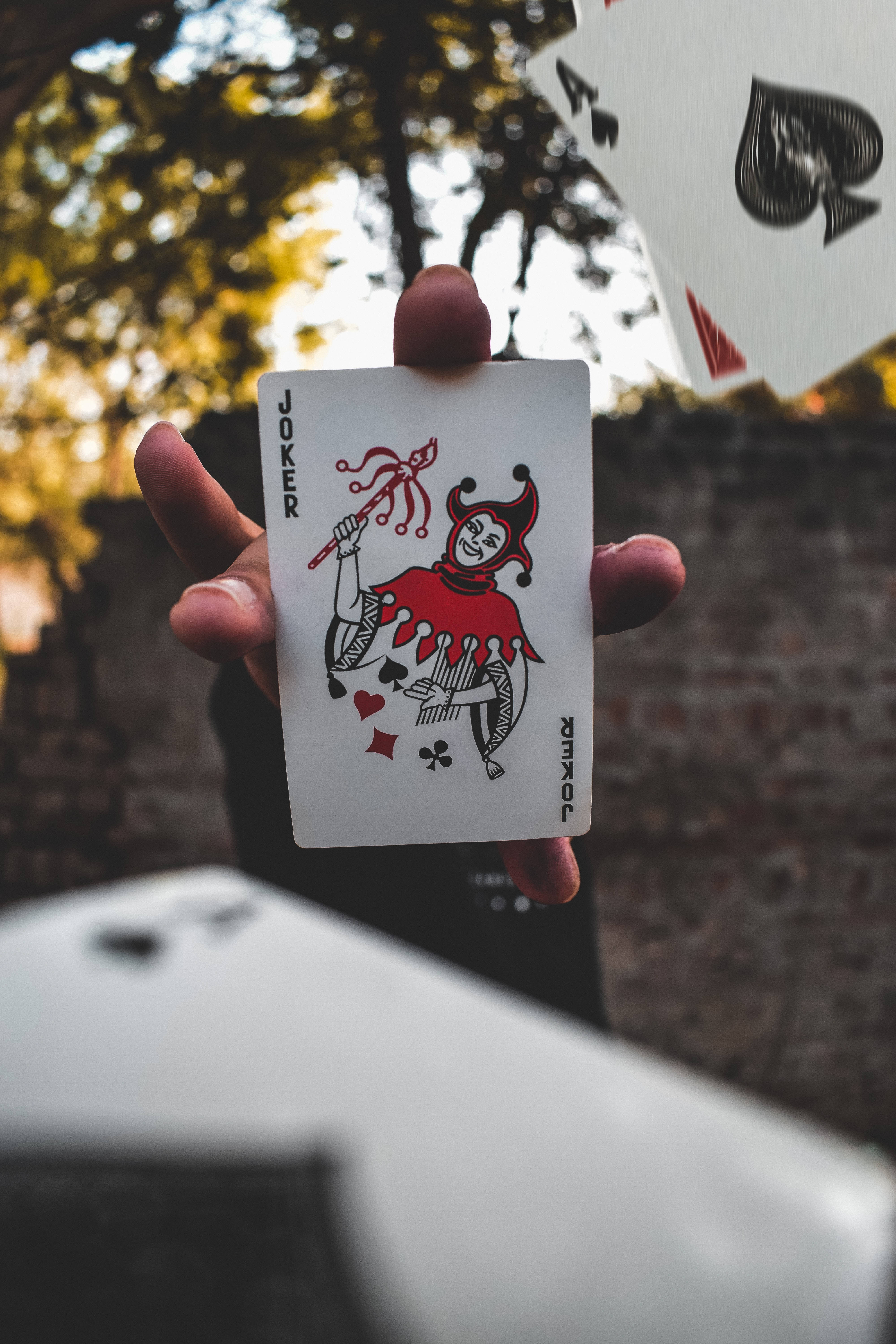 The Joker - The Ultimate Wild Card 壁紙 — 一隻手喺戶外揸住一張紅色嘅小丑啤牌，背景有秋天嘅樹同磚牆，呈現柔和焦點。