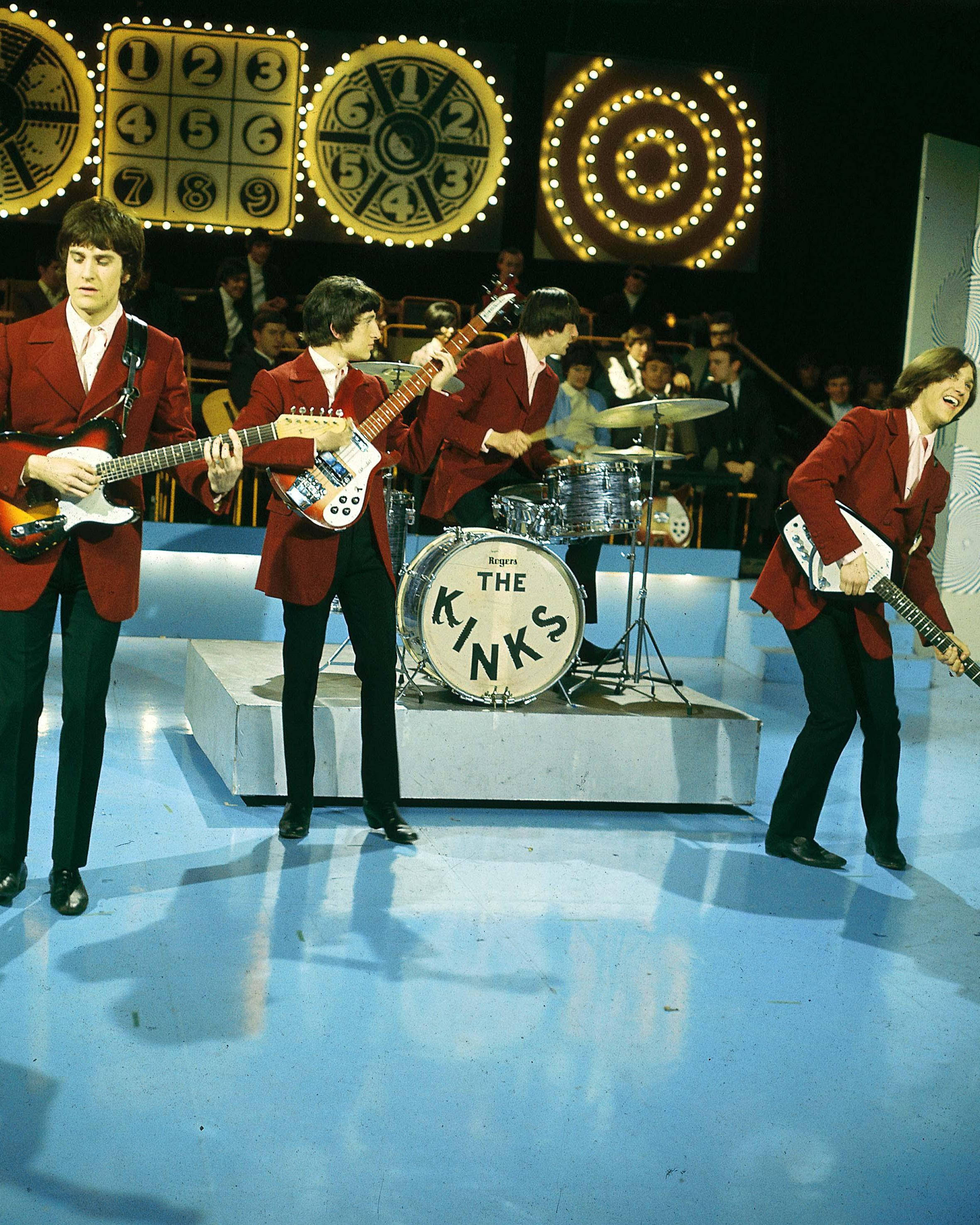 The Kinks 1965現場演出海報 壁紙 — The Kinks 在充滿活力的藍色舞台上表演，在發光的時鐘主題燈光下穿著相配的紅色外套。