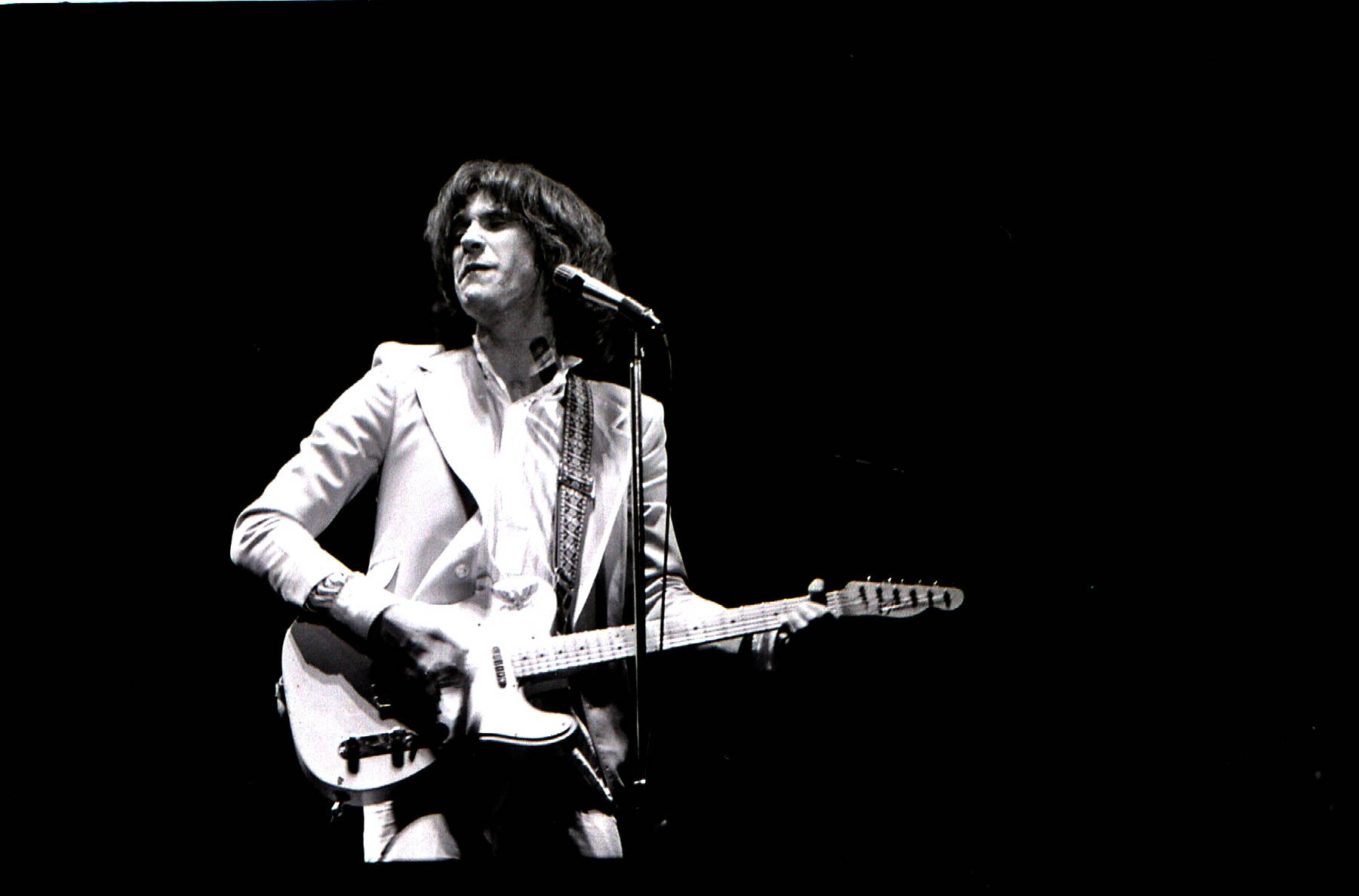 The Kinks Ray Davies 海報 壁紙 — 一位身穿白色西裝的音樂家在黑色背景的舞台燈光下熱情地彈奏電結他。