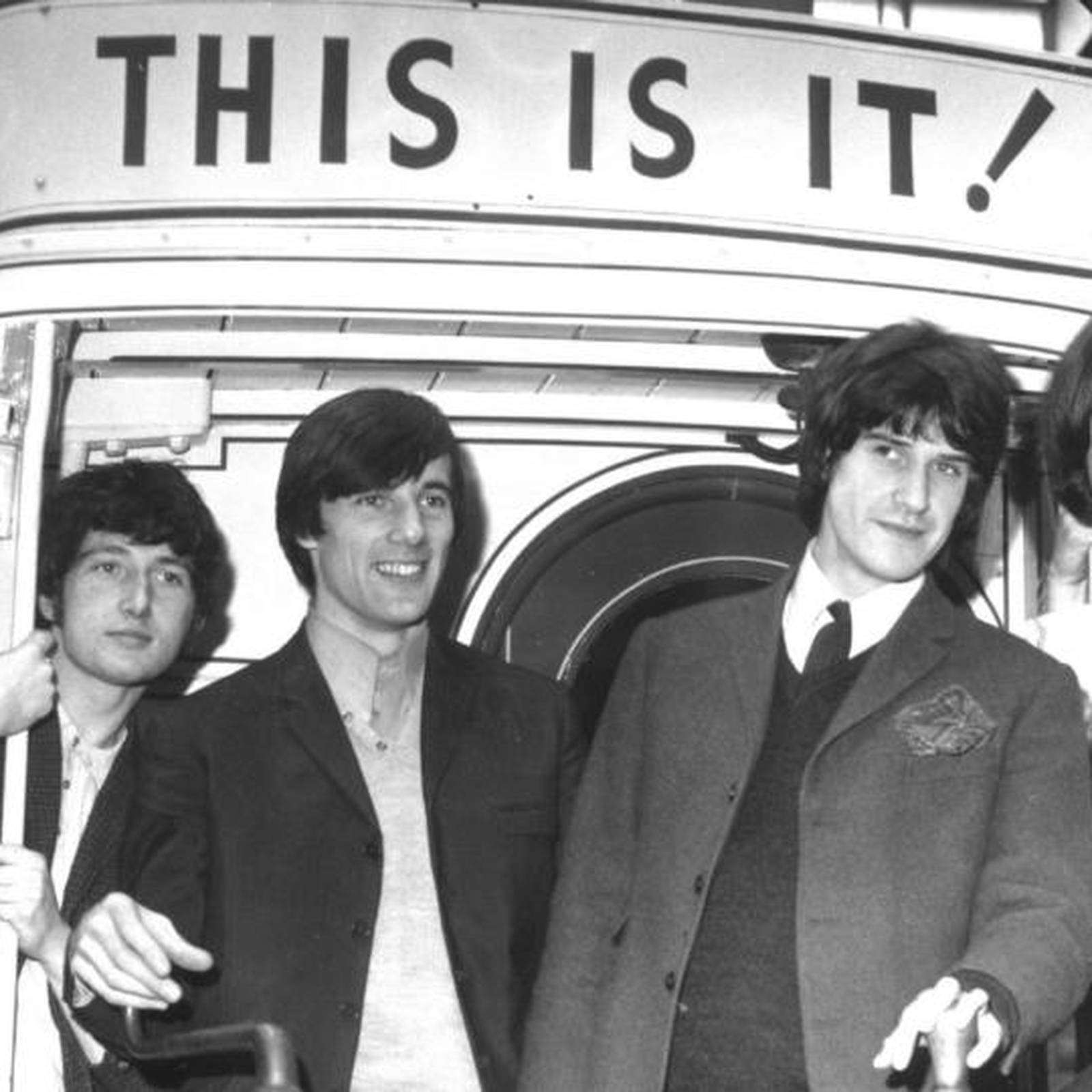 The Kinks「This Is It!」海報 壁紙 — 三位身穿西裝的男士微笑站在一輛巴士旁，巴士上方有一個醒目的 'THIS IS IT!' 標誌，是一張經典黑白照片。