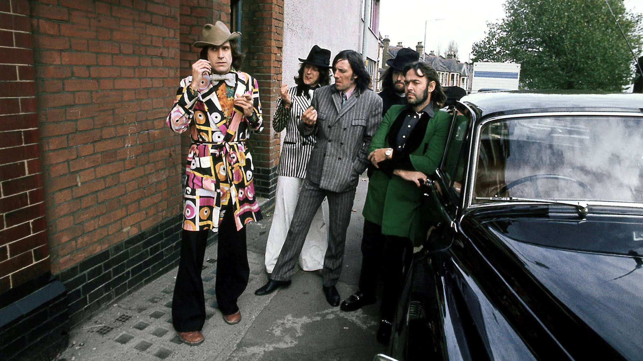 The Kinks 复古街頭照片 壁紙 — 一群穿著大膽1970年代時尚的男士站在磚砌人行道旁的一輛復古黑色汽車旁邊。