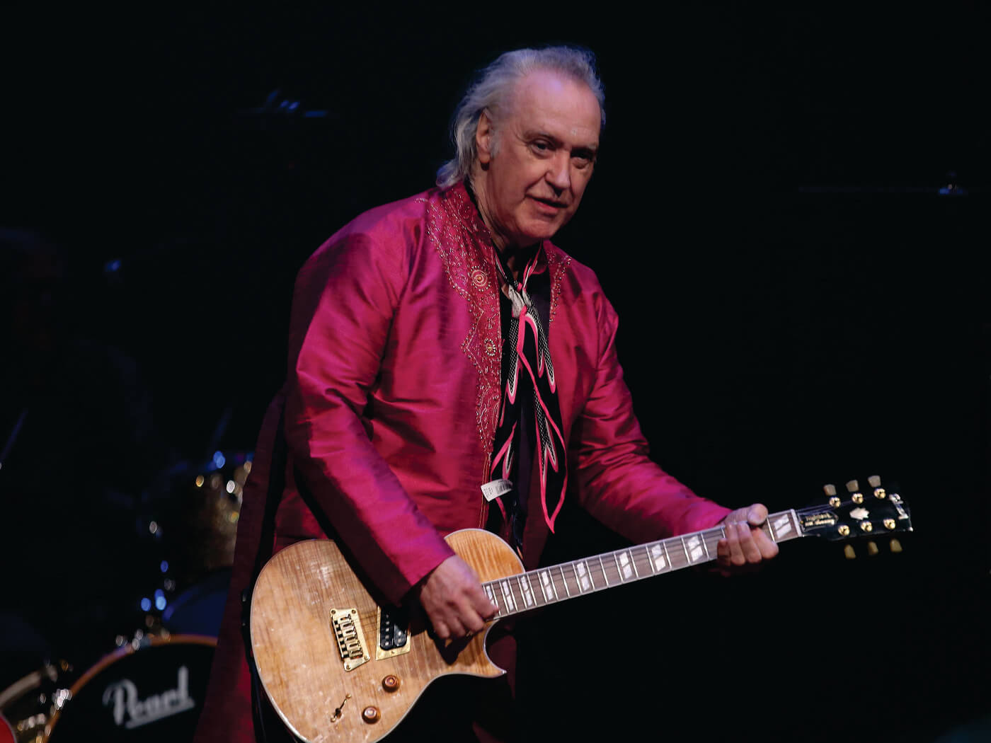 The Kinks 的老 Dave Davies 壁紙 — 一位身穿鮮紅色外套的音樂家在舞台燈光下彈奏淺色木電結他，身後可見一套鼓。