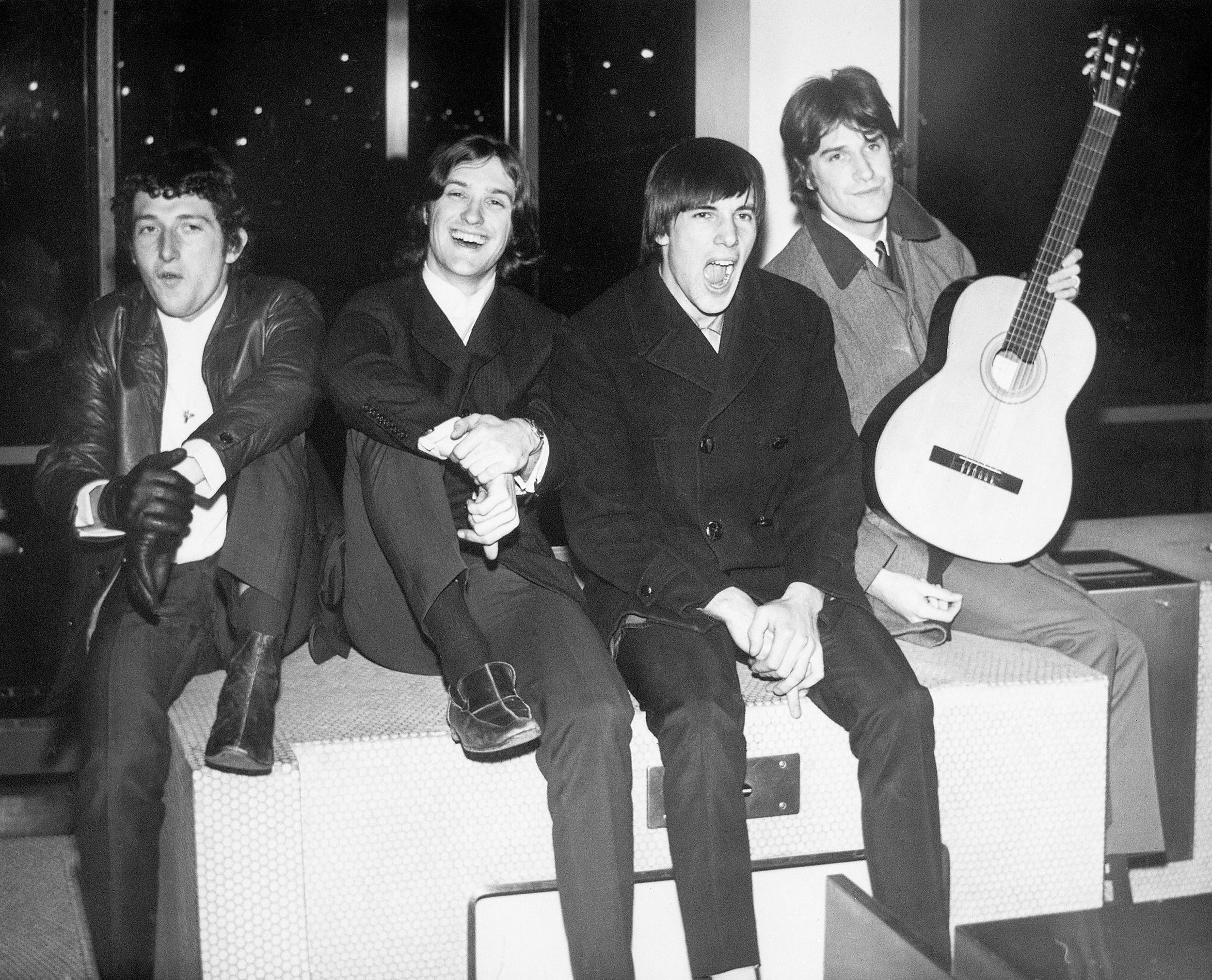 The Kinks - 經典搖滾樂隊復古肖像 壁紙 — 四位身穿1960年代服裝的男子坐在現代長椅上，其中一人手持古典結他，所有人以富有表情的面孔擺姿勢，黑白照片。