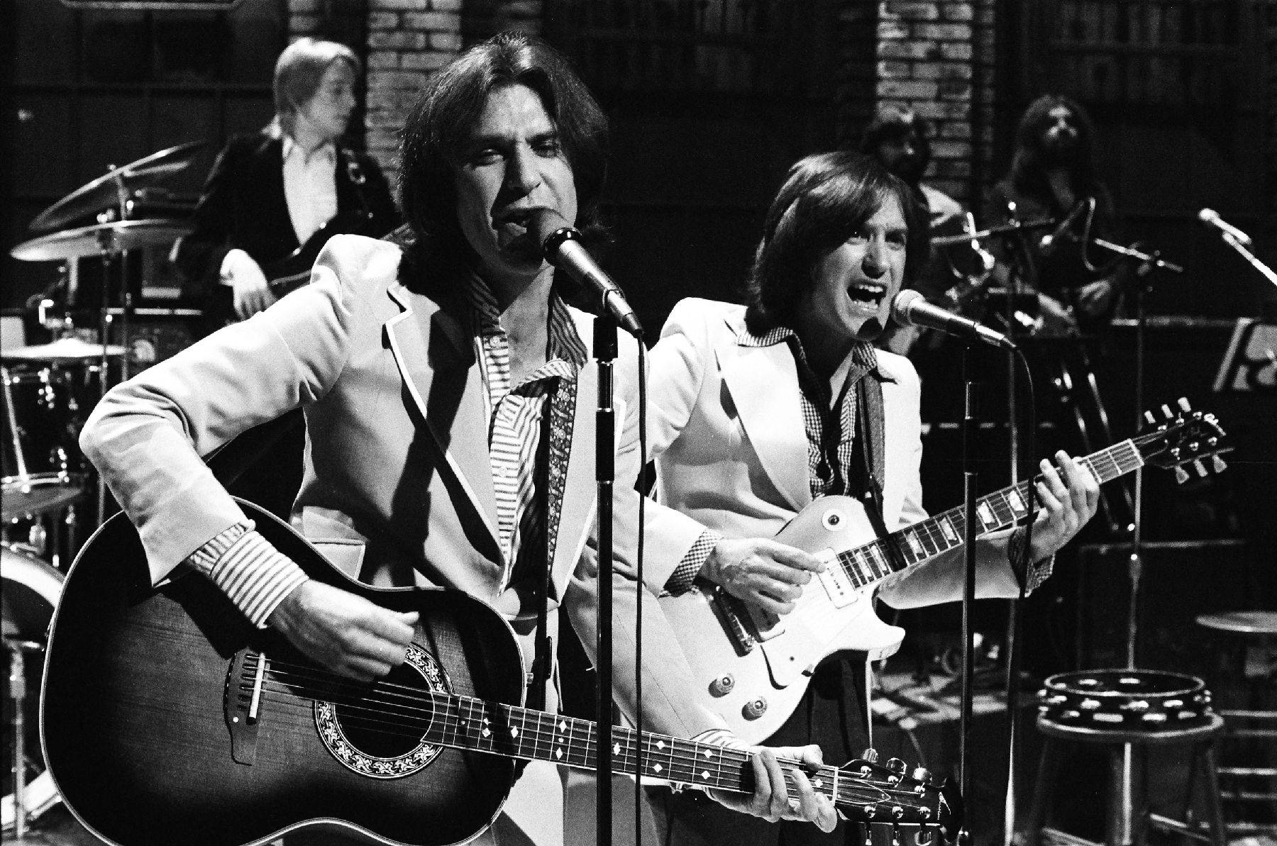The Kinks 黑白現場演出 壁紙 — 兩位身穿白色西裝的音樂家在戲劇性的舞台燈光下，帶著結他和咪高峰在舞台上熱情演出。
