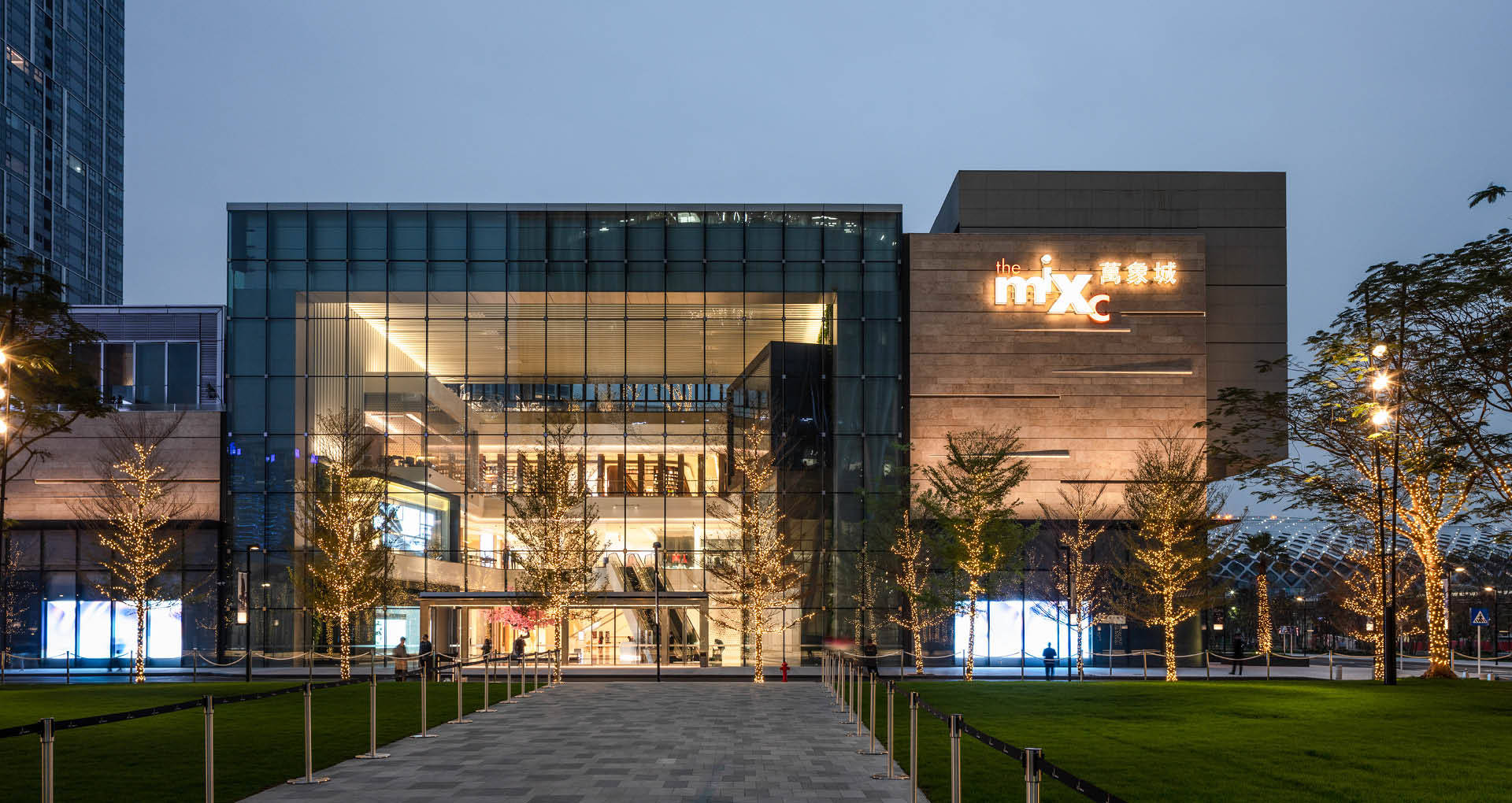 深圳The MIXC 壁紙 — 現代玻璃和石材建築在黃昏時亮起，發光的樹木和鋪砌的人行道通往入口。