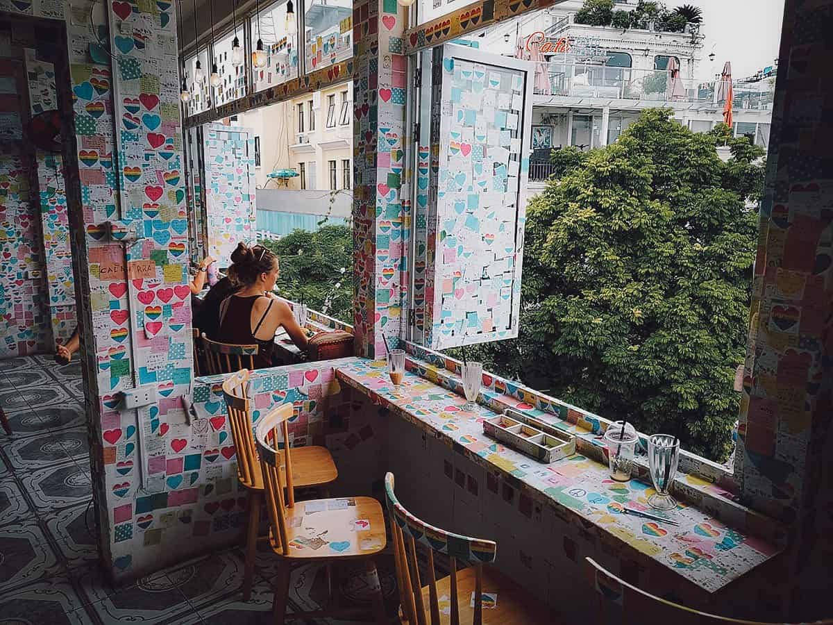 河內The Note咖啡館 壁紙 — 一位女士坐在色彩繽紛、裝飾著心形的窗邊座位，身處充滿活力的咖啡館，窗外是茂密的綠樹和城市建築。