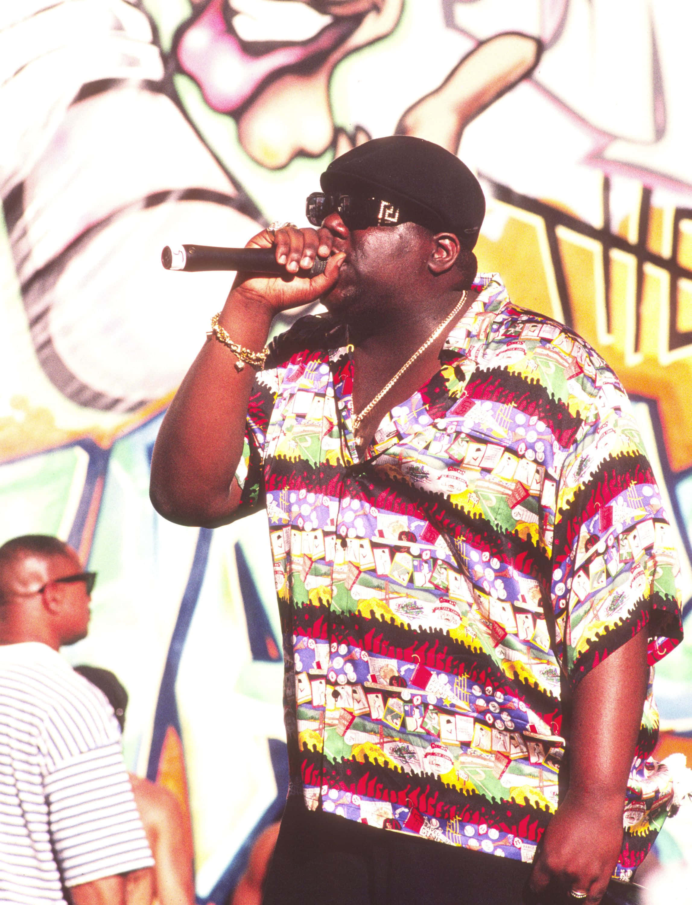 The Notorious Big 在 1995 年 Beat Summer Jam 演唱會上的風采 壁紙 — 一位身穿鮮豔圖案襯衫和黑色帽子的說唱歌手，在色彩繽紛的塗鴉背景下對著麥克風表演。