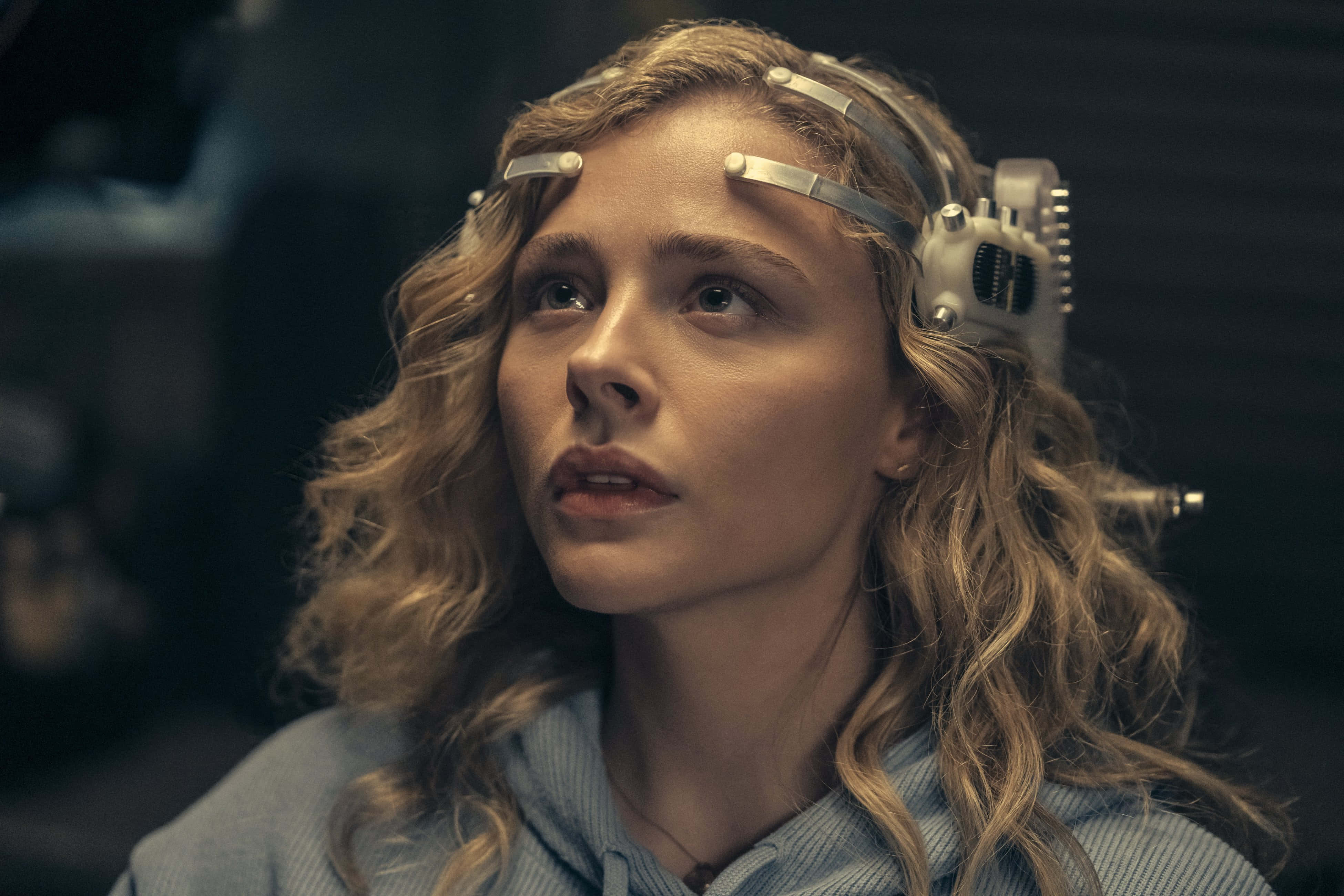 《The Peripheral》劇集中Flynne Fisher頭戴的奇特科技裝置 壁紙 — 一位年輕女性，金色捲髮，戴著帶有金屬感應器的未來感頭帶，在昏暗的房間裡向上凝視。
