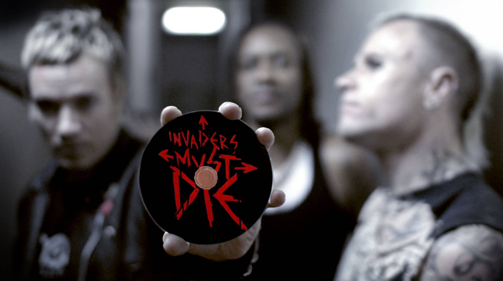 手拿著一張黑色CD，上面有紅色文字「Invaders Must Die」，前面是模糊的樂隊。
