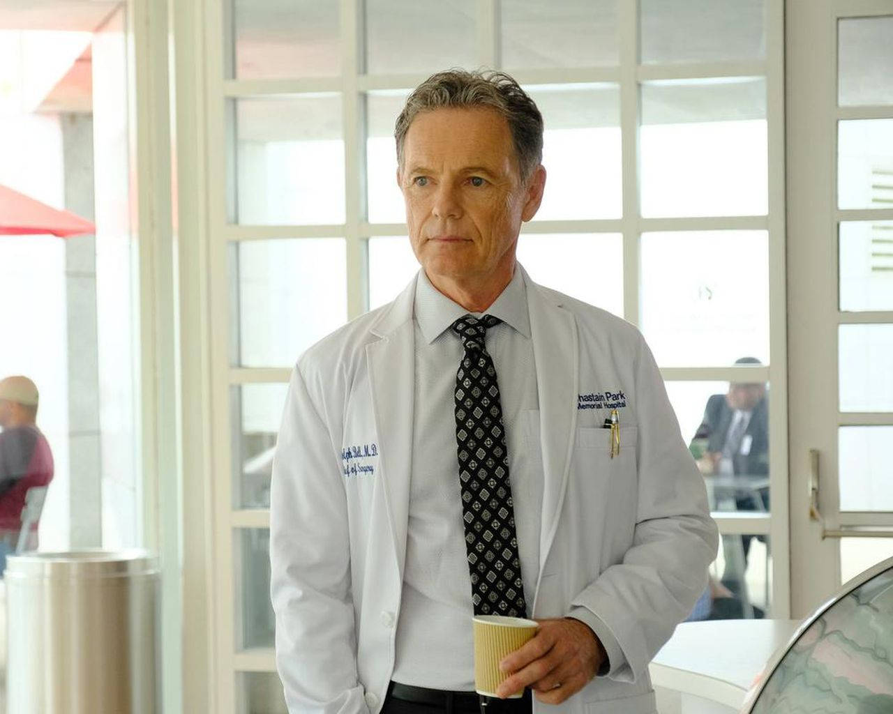《The Resident》劇中醫生貝爾布魯斯·格林伍德 壁紙 — 一位嚴肅的中年醫生穿著白色實驗室外套，在明亮的醫院大廳裡拿著一杯咖啡。