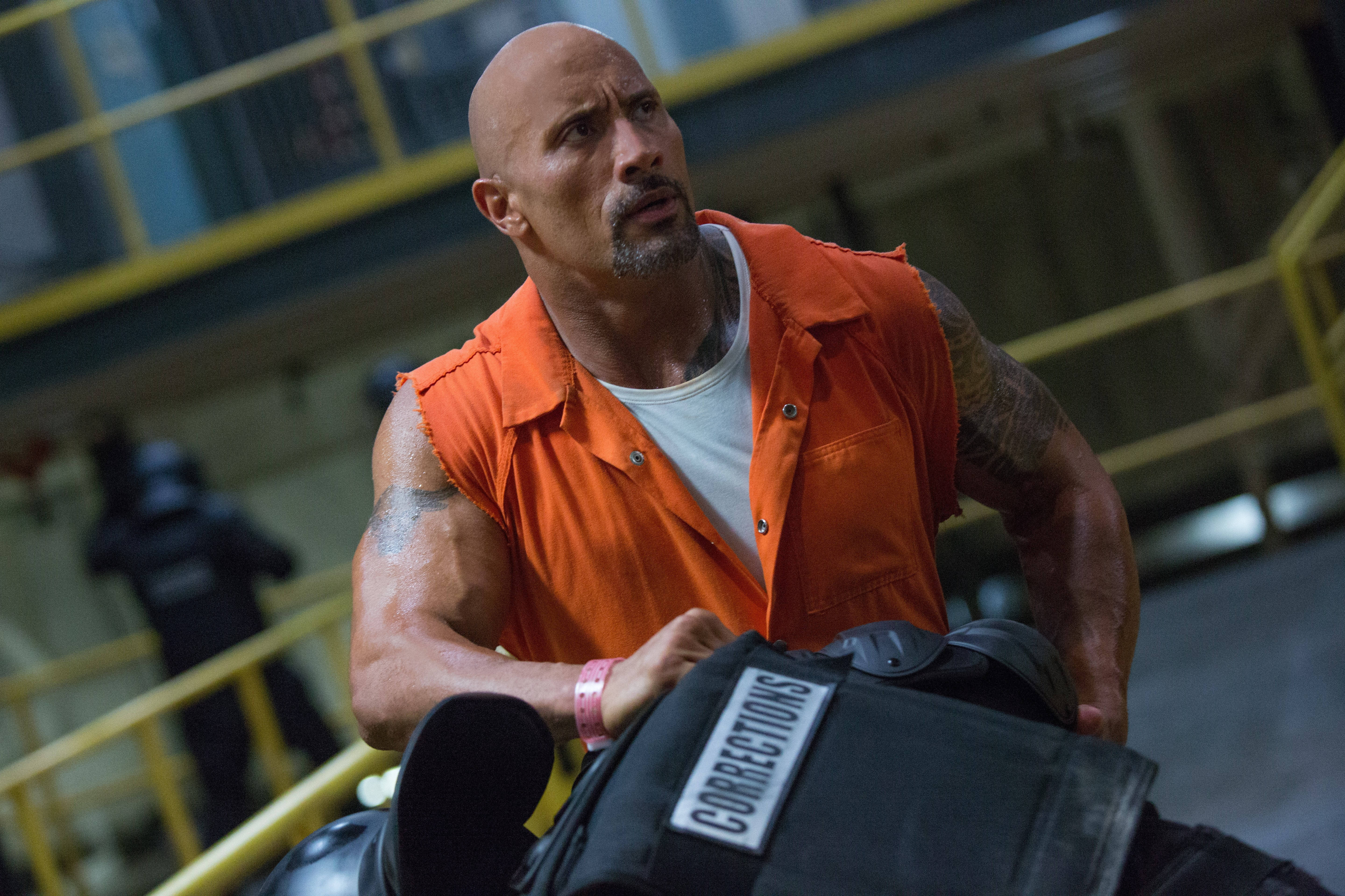 The Rock 穿囚服造型 壁紙 — 狄維·莊遜身穿橙色監獄連身衣，在緊張的工業環境中抓住懲教人員的背心。