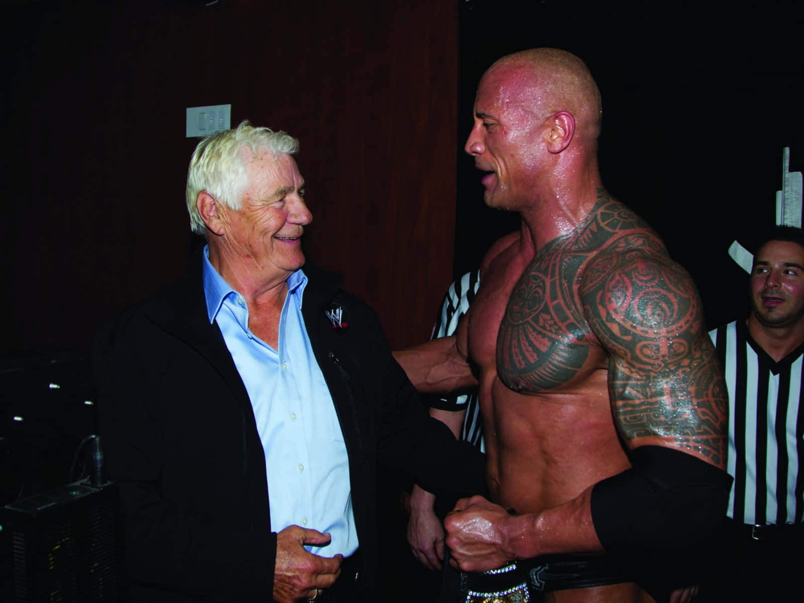 帕特·帕特森與 The Rock 會面 壁紙 — 一位身穿黑色外套的年長男性在燈光昏暗的後台區域，對一位肌肉發達、滿身紋身的摔角手露出溫暖的笑容。