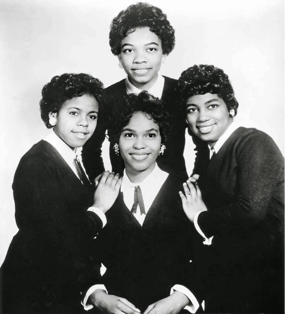 The Shirelles 1970年經典合照 壁紙 — 四位微笑的女性穿著相配的深色西裝，在簡單的攝影棚背景前緊密地擺姿勢，呈現經典的黑白肖像。