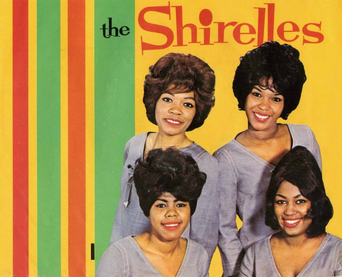 The Shirelles《Don't Say Goodnight And Mean Goodbye》黑膠專輯封面 壁紙 — 四位微笑的女性穿著相配的灰色上衣，在充滿活力的黃、綠、紅條紋背景前擺姿勢，背景上有粗體紅色的「the Shirelles」字樣。