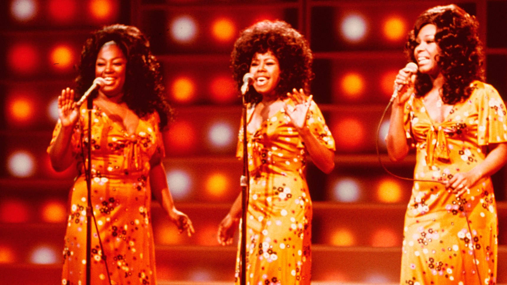 The Shirelles《Midnight Special》1970 節目演出 壁紙 — 三位微笑的女性穿著相配的花卉連衣裙，在溫暖照明的舞台上表演，身後有發光的圓形燈光。