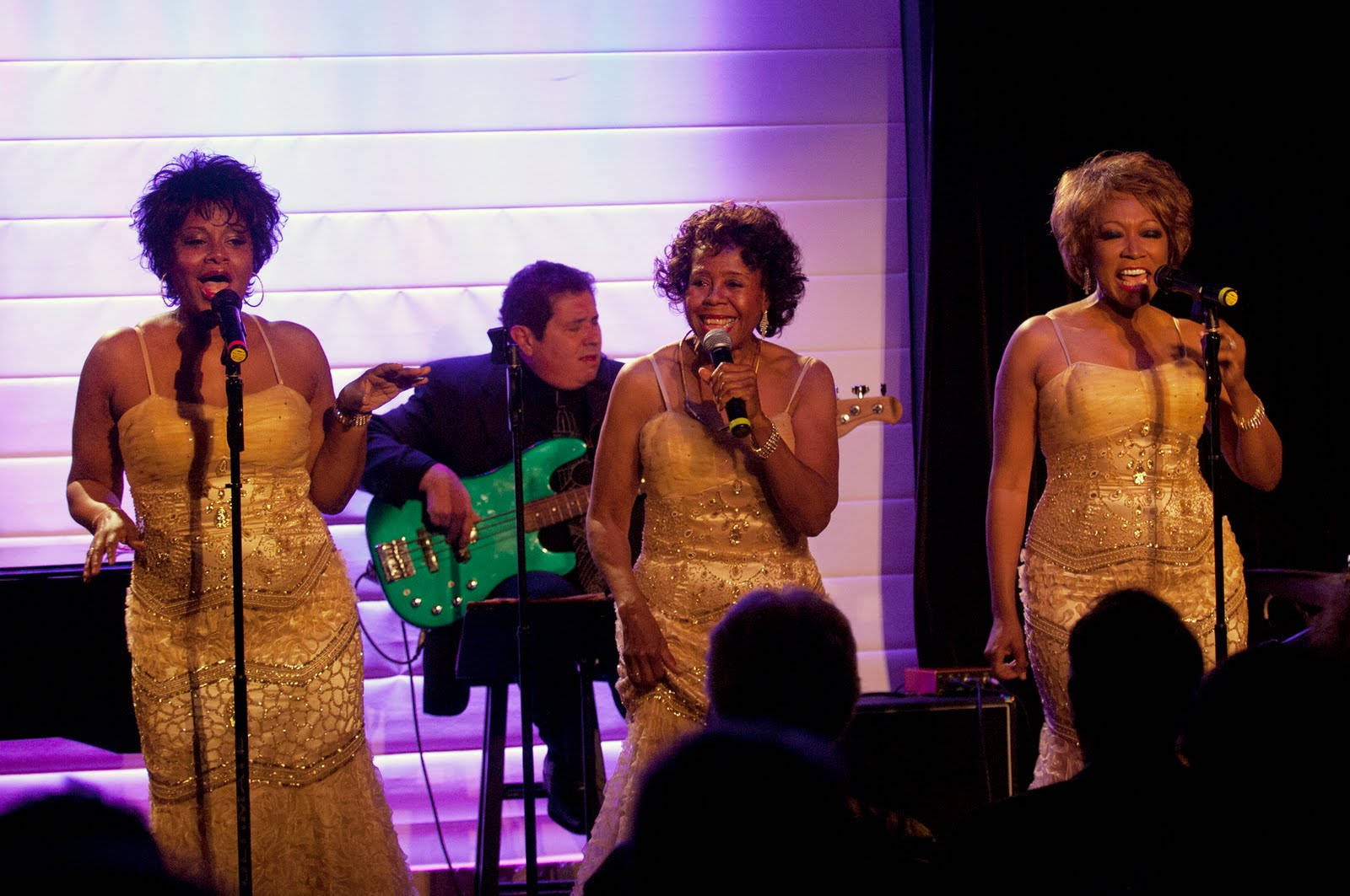 The Shirelles 女子組合演唱會現場 壁紙 — 三位身穿閃亮金色連衣裙的女性在燈光璀璨的舞台上對著麥克風唱歌，身後有一位貝斯手。