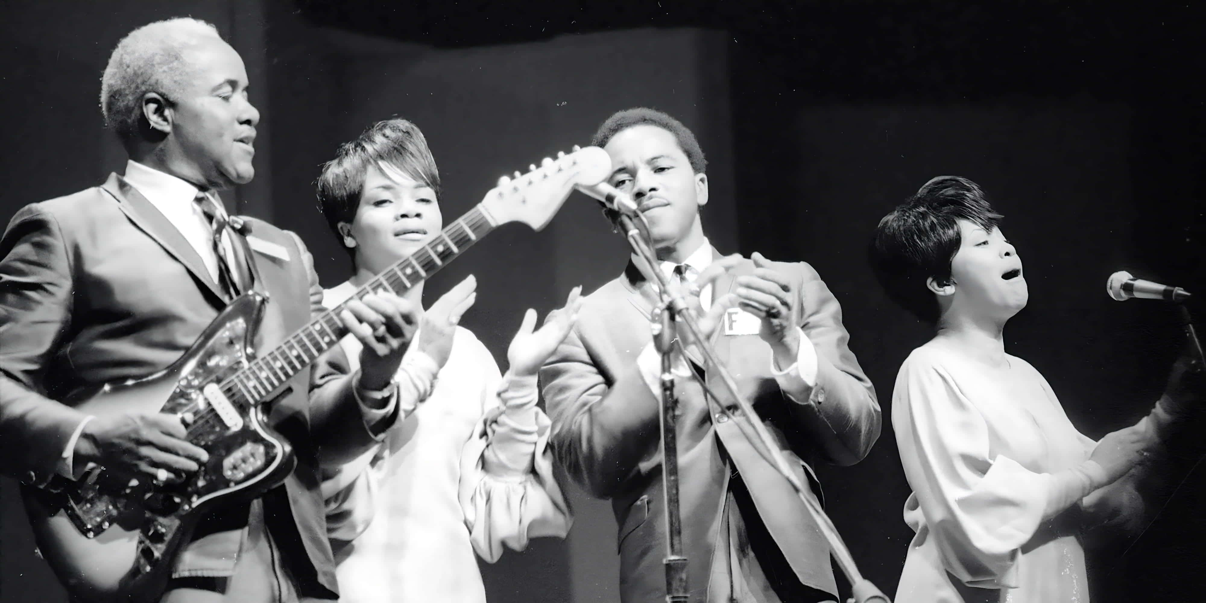 The Staple Singers 1967年現場演出 壁紙 — 一張黑白照片，四位音樂家在舞台上表演，有結他手、歌手和咪高峰，在戲劇性的燈光下。