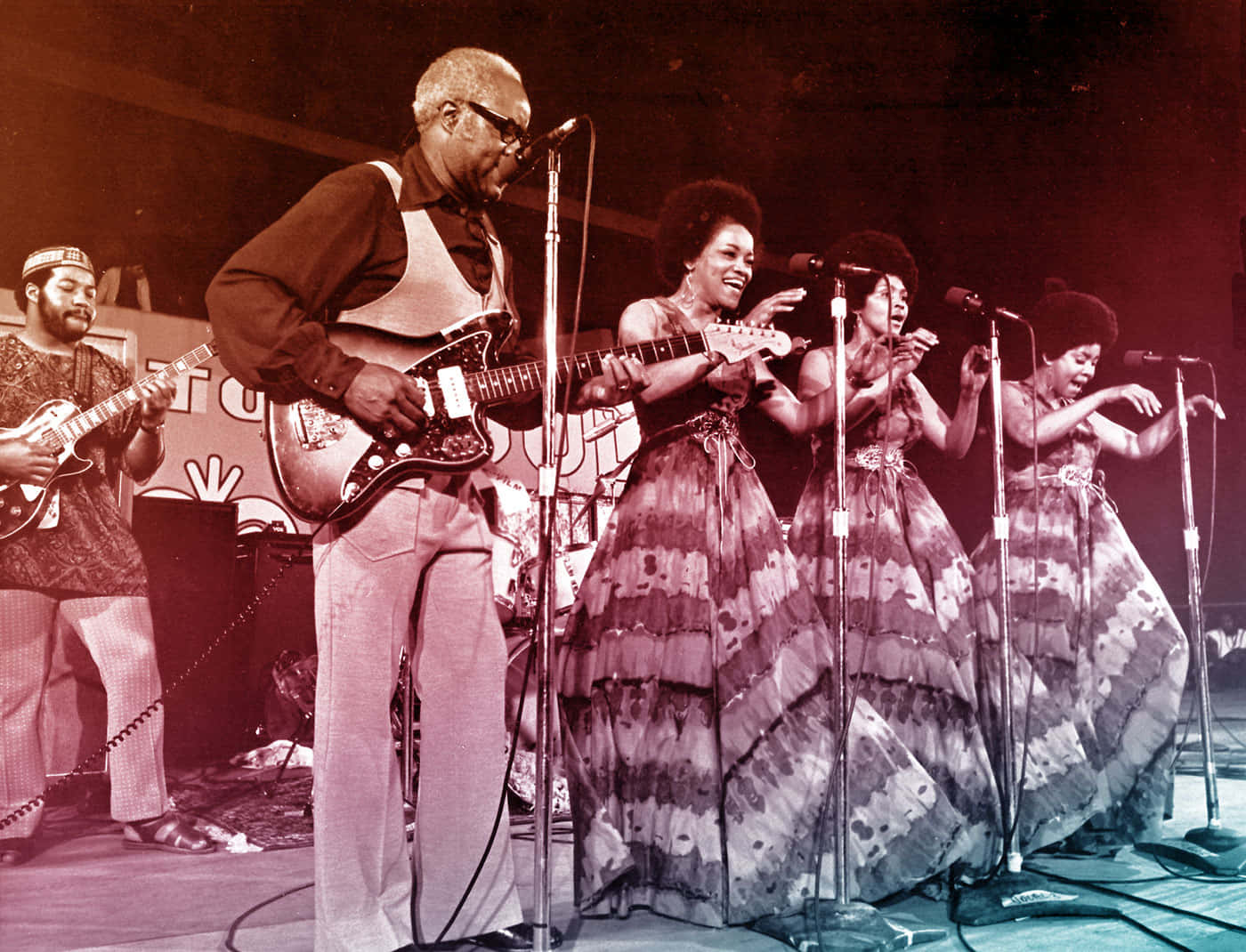 The Staple Singers 1971年加納現場演出 壁紙 — 一張復古棕褐色調的照片，拍攝了一支樂隊在舞台上表演，有一位結他手和四位穿著飄逸圖案連衣裙的歌手。