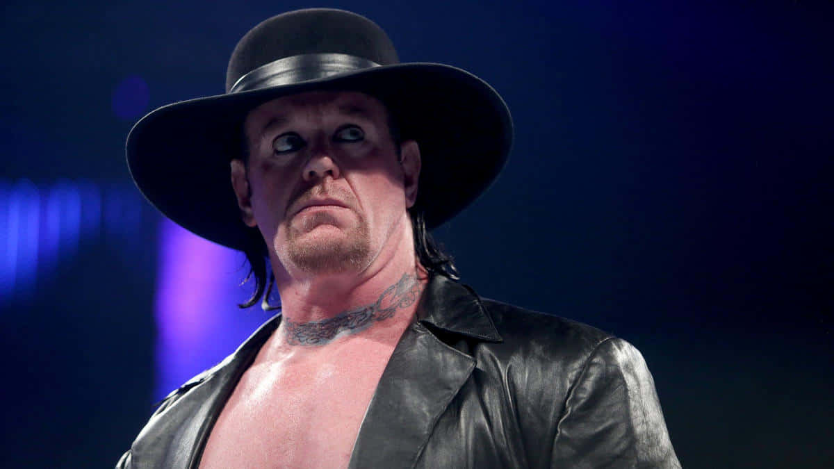  壁紙 — The Undertaker 戴著標誌性的黑色帽子和皮褸，在戲劇性的藍色舞台燈光下凝視著。